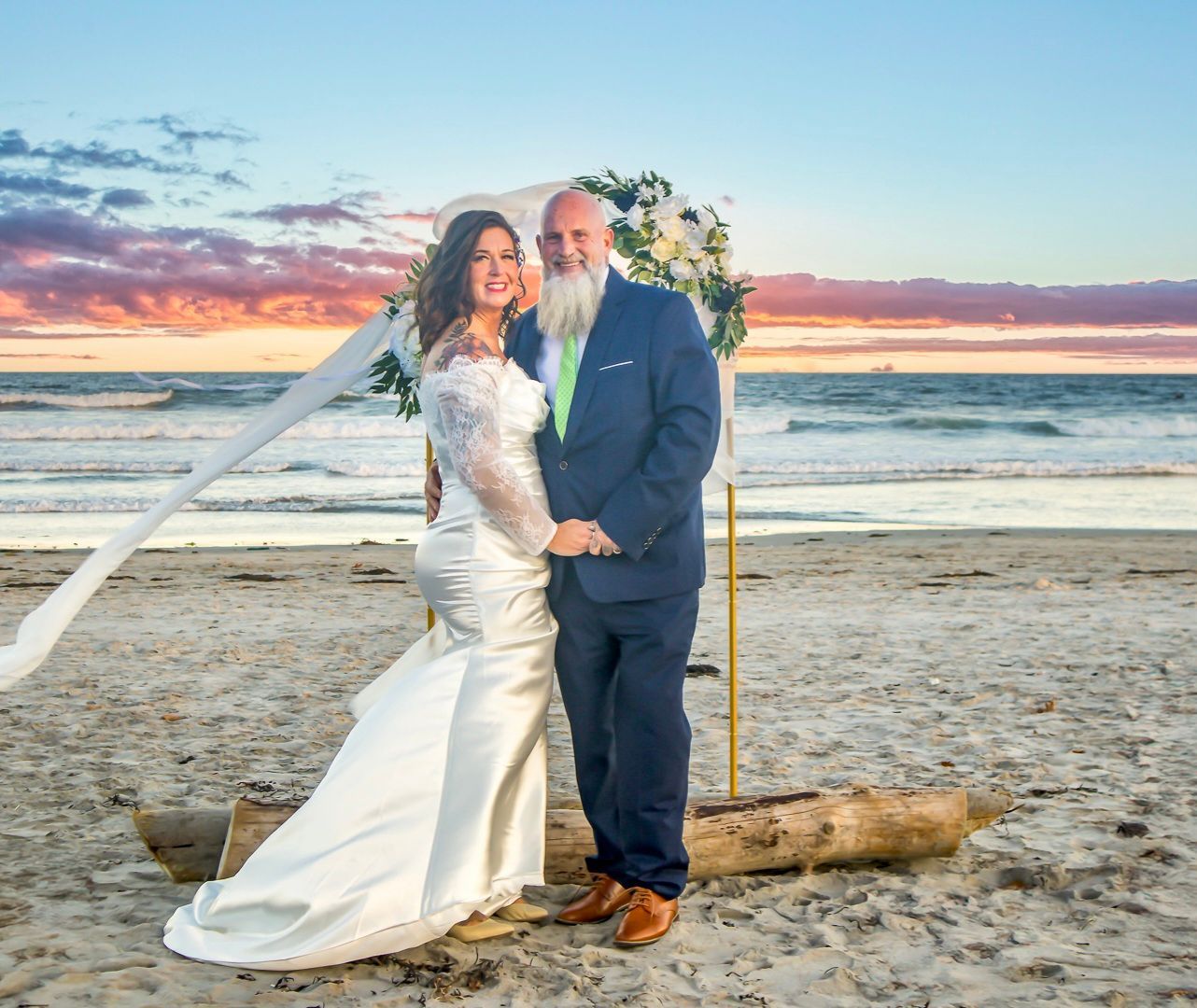 Wedding couple posing on a beach at sunset, with ocean waves and pink clouds behind them.