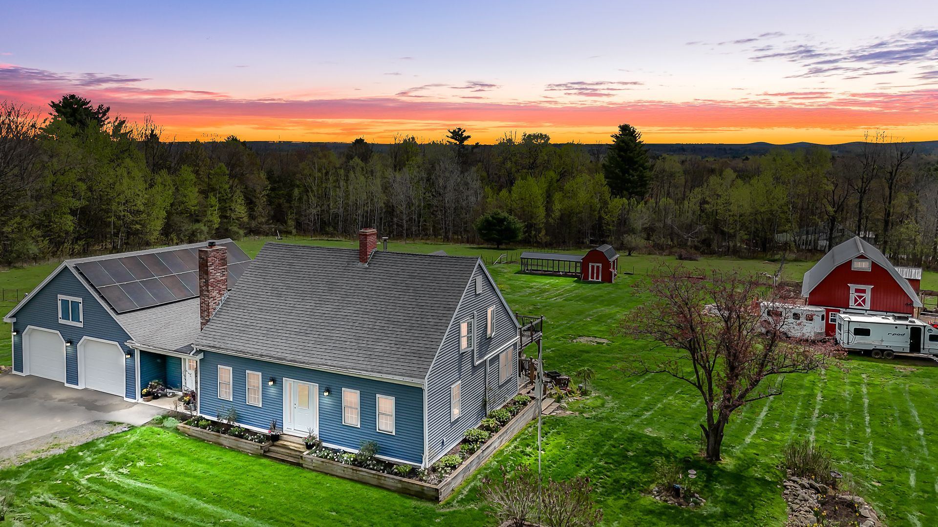 Blue farmhouse and red barn in a green rural yard at sunset with a pink-orange sky