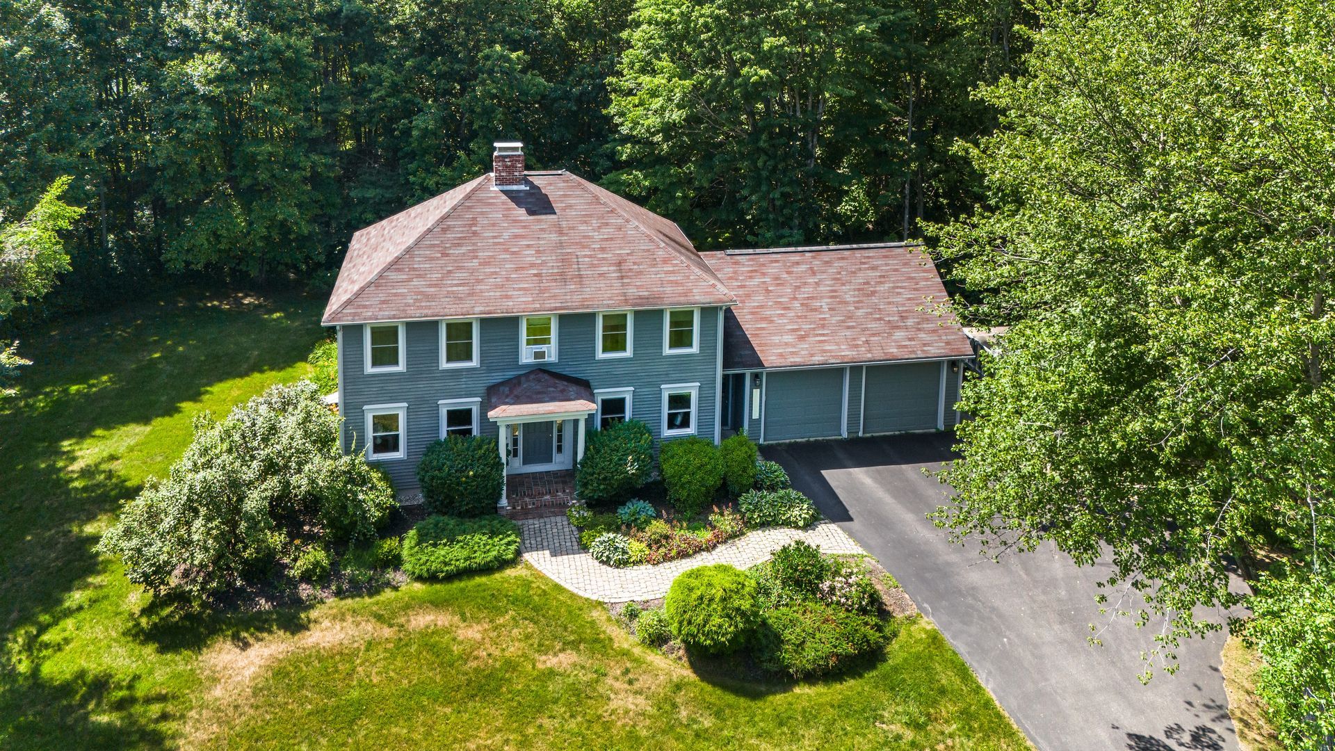 Aerial view of a gray two-story house with a driveway, surrounded by green trees and lawn.