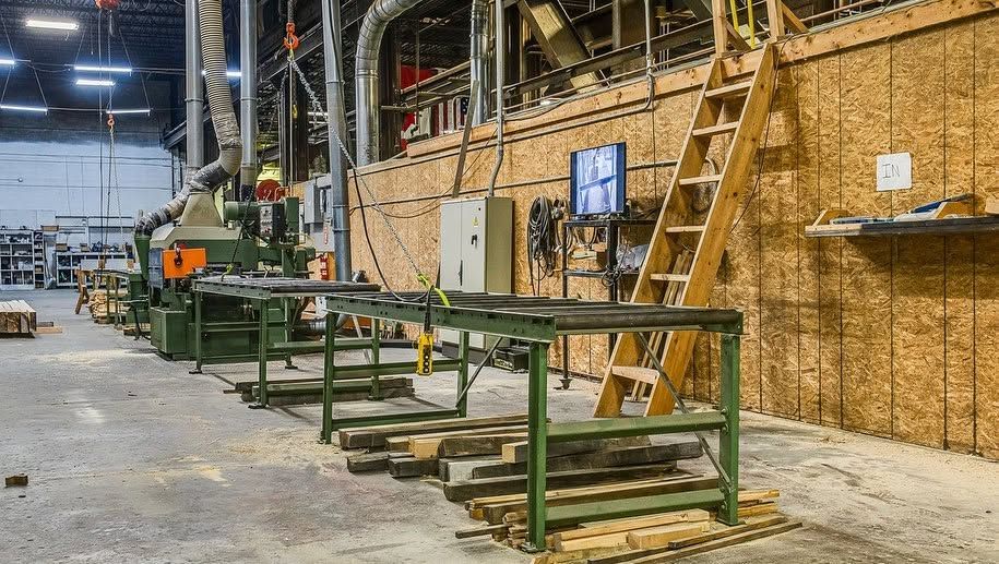 Industrial warehouse with green workbenches, ladders, and shelving against a plywood wall