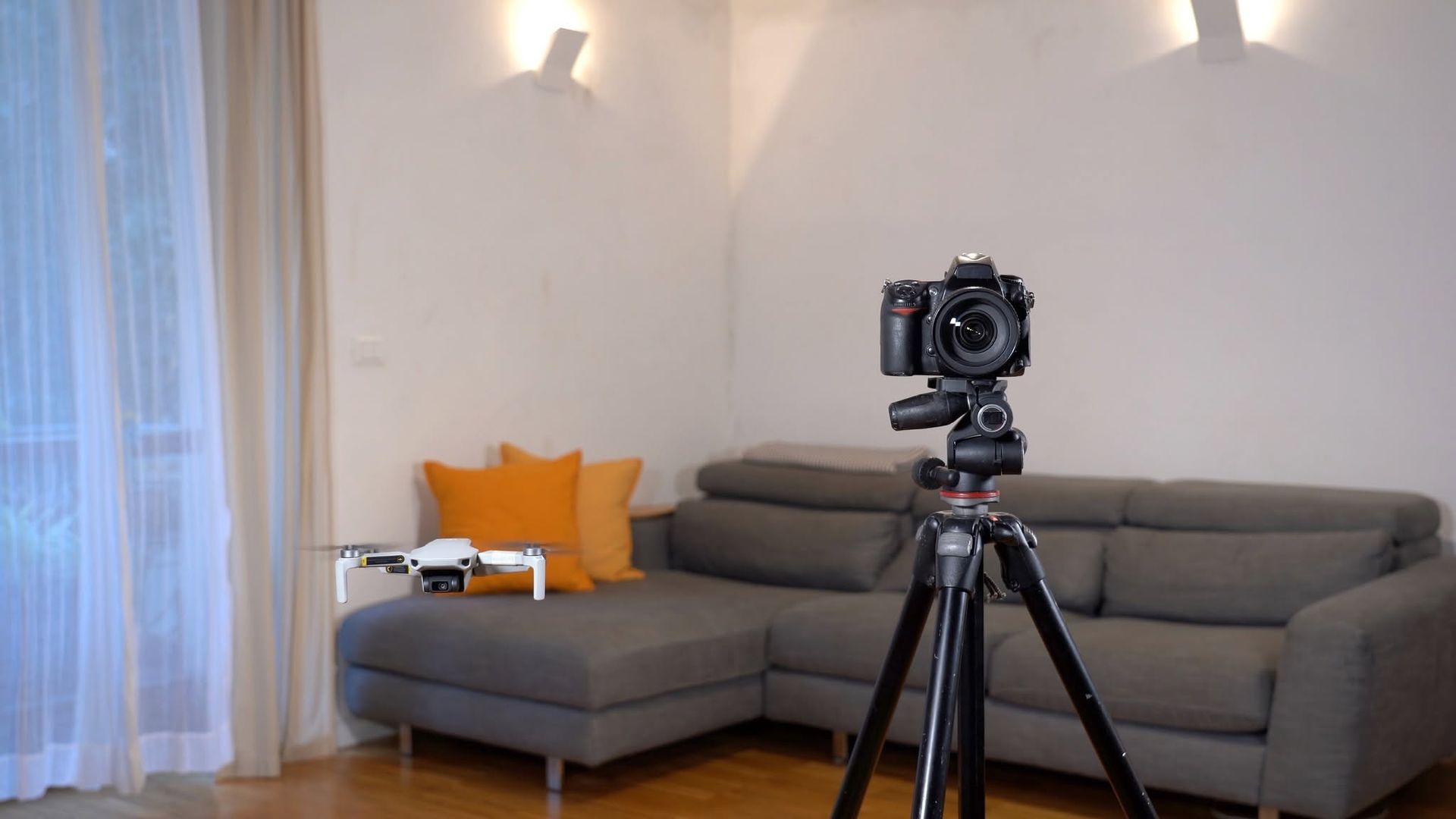 Camera on a tripod in a modern living room with a gray sofa, orange pillows, and sheer curtains.