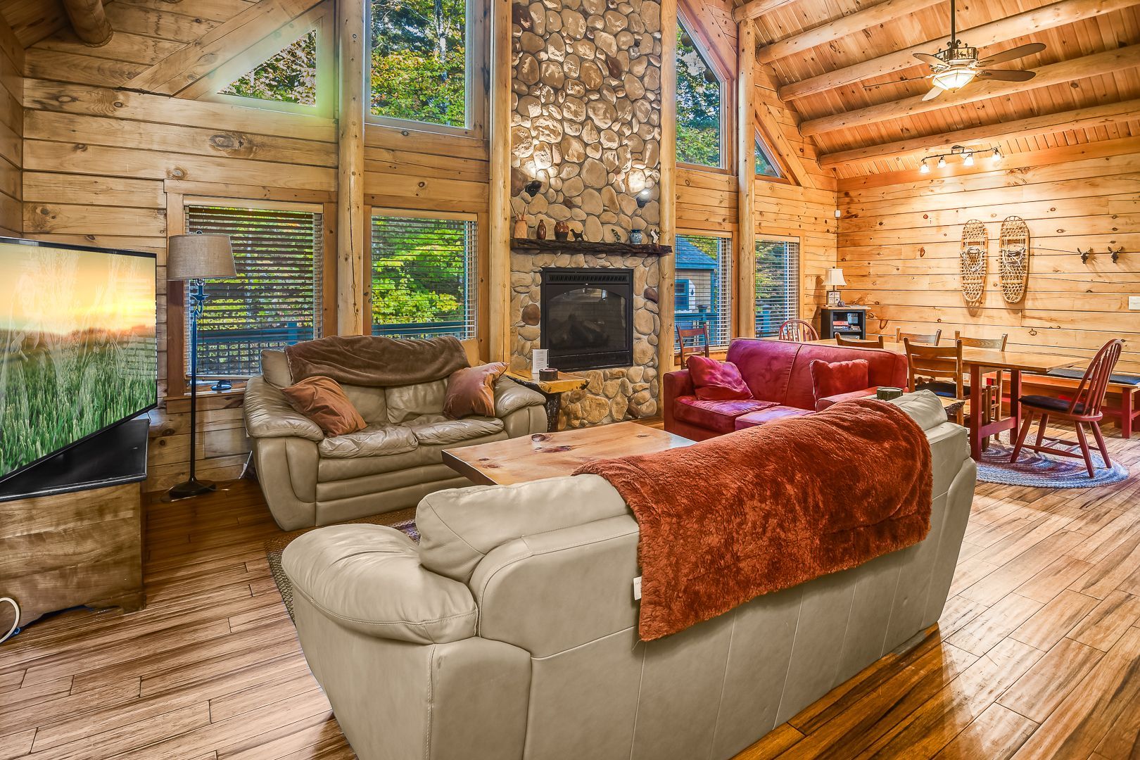 Rustic log cabin living room with stone fireplace, leather sofas, and large windows overlooking trees