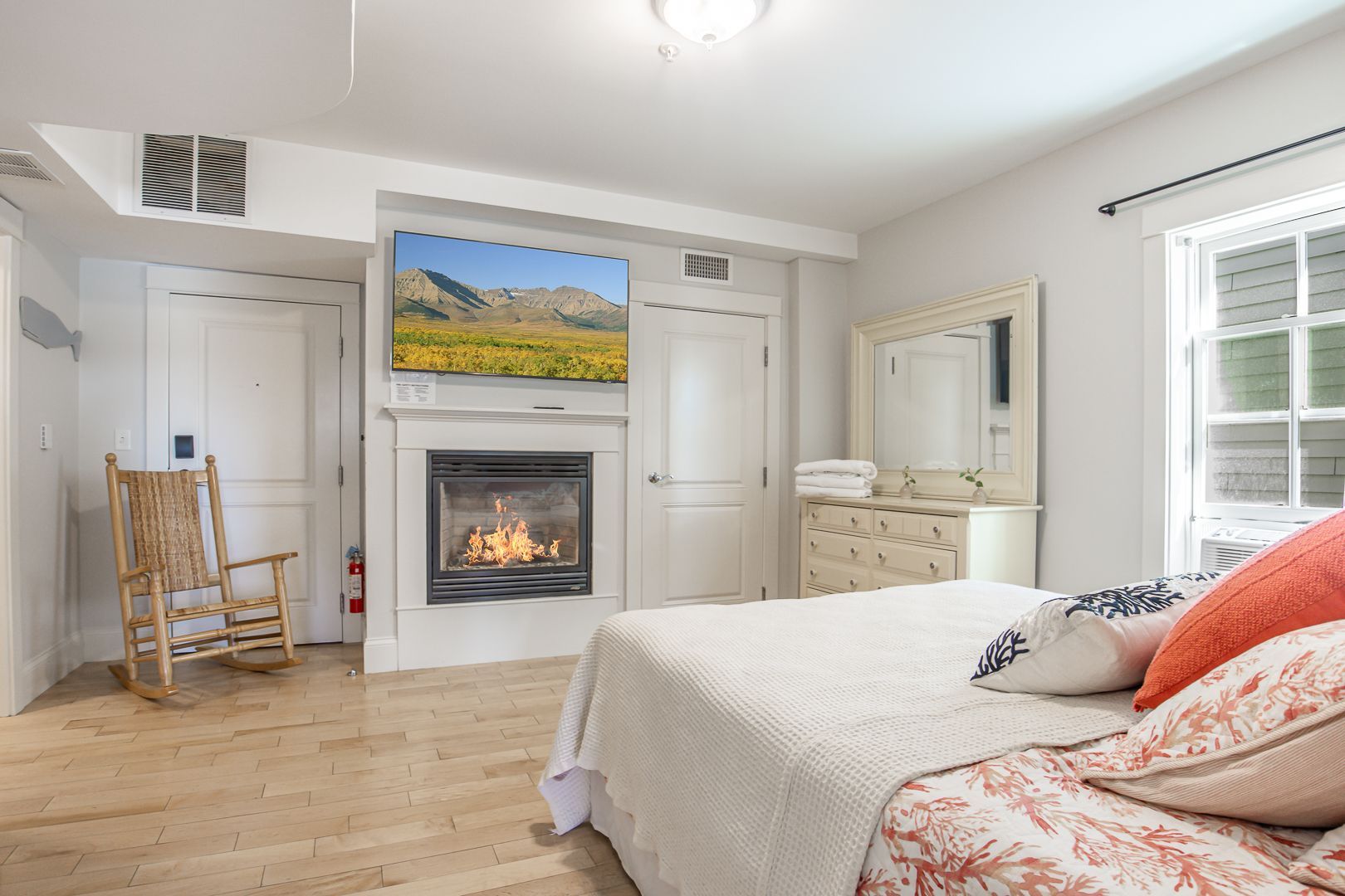 Bright bedroom with fireplace, white dresser, bed, and yellow rocking chairs by a window