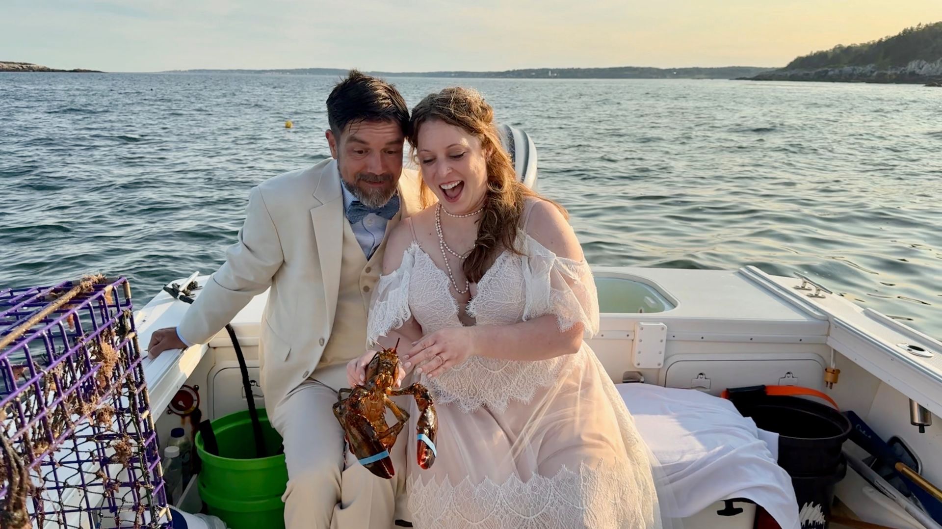 Couple in white outfits smiling on a boat at sunset, with calm water behind them.