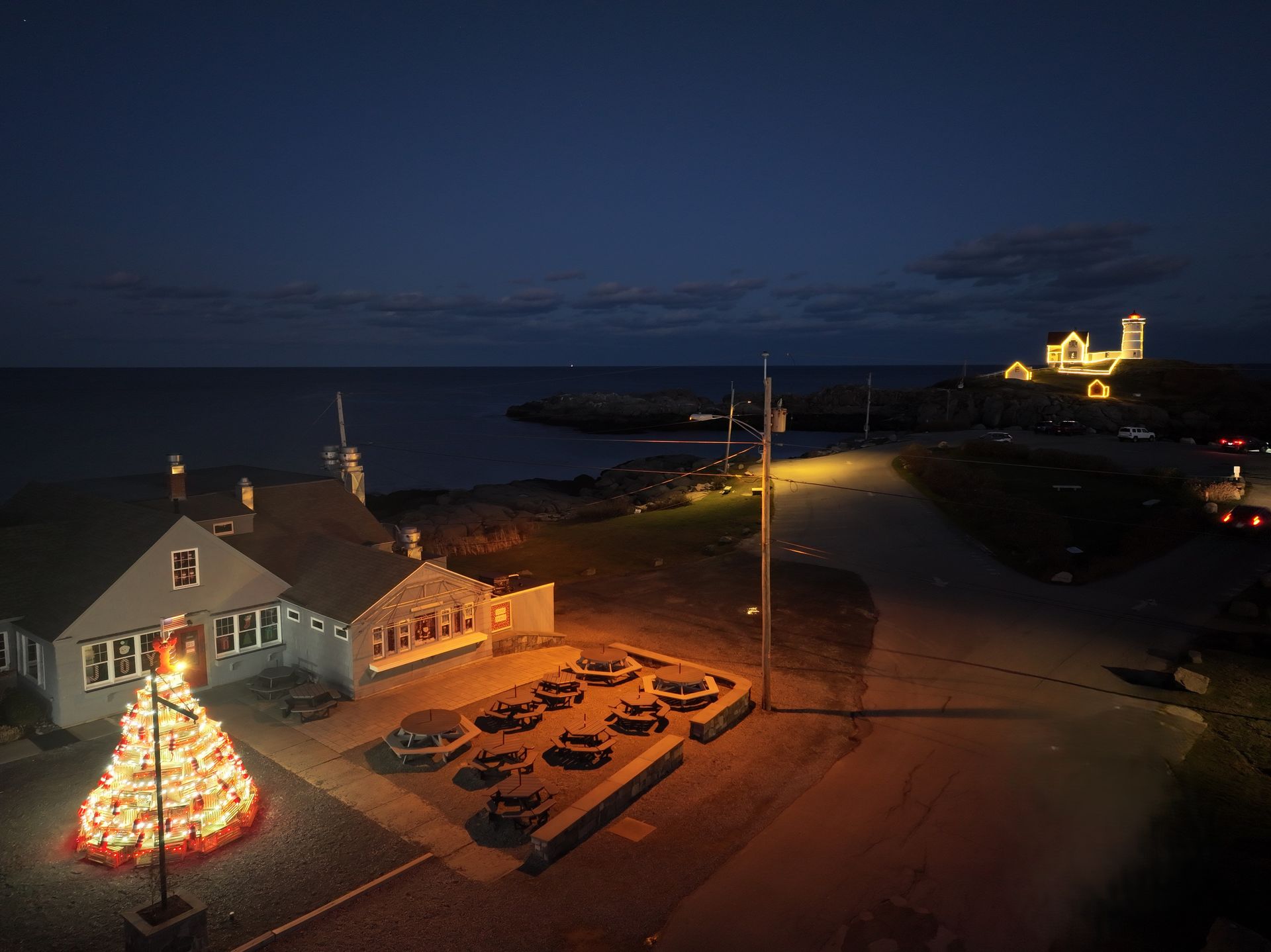 Nighttime harbor scene with a lit seaside house, parked cars, and a brightly illuminated building on the shoreline