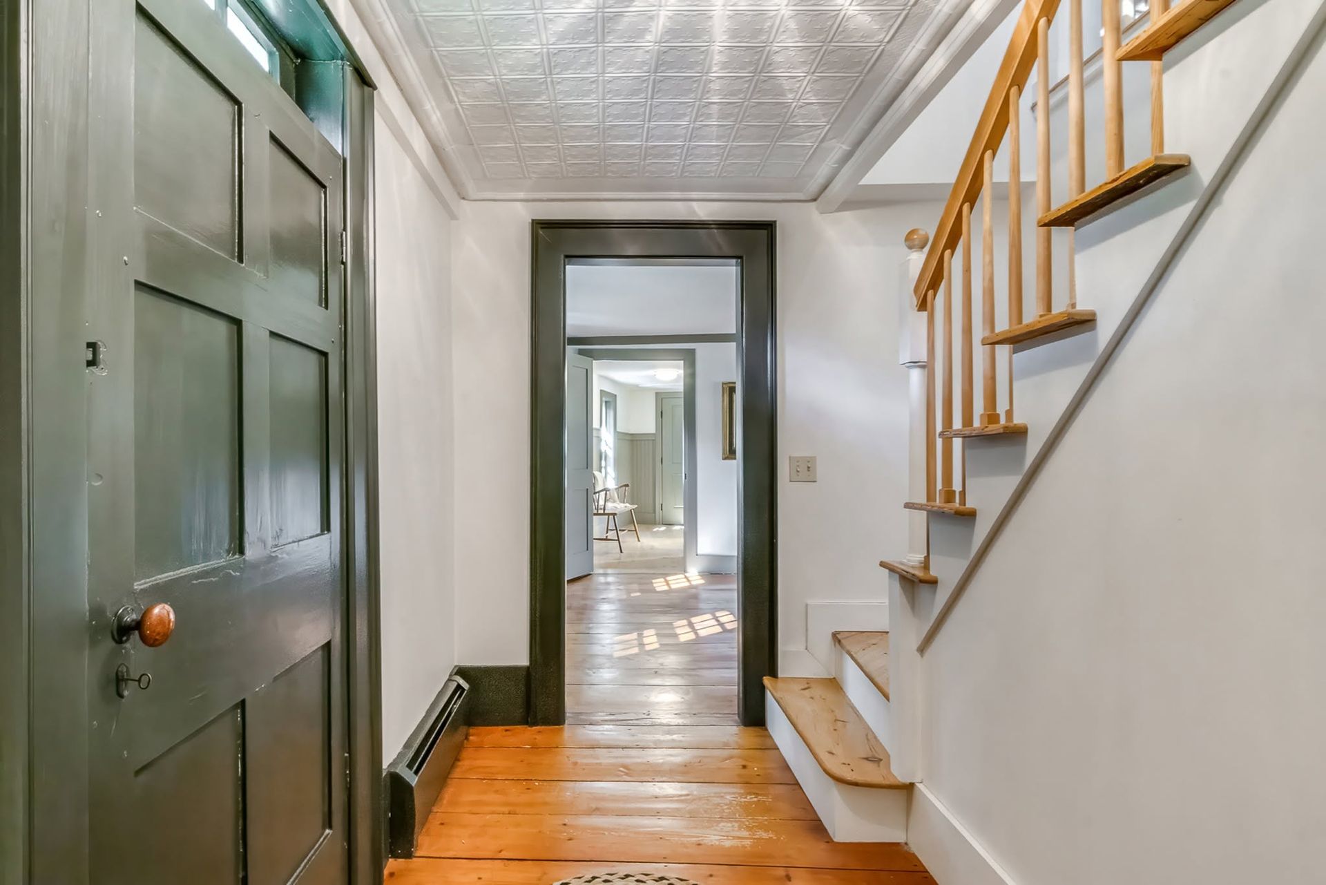Narrow hallway with wood floors, green door, staircase, and a view into a brighter room beyond