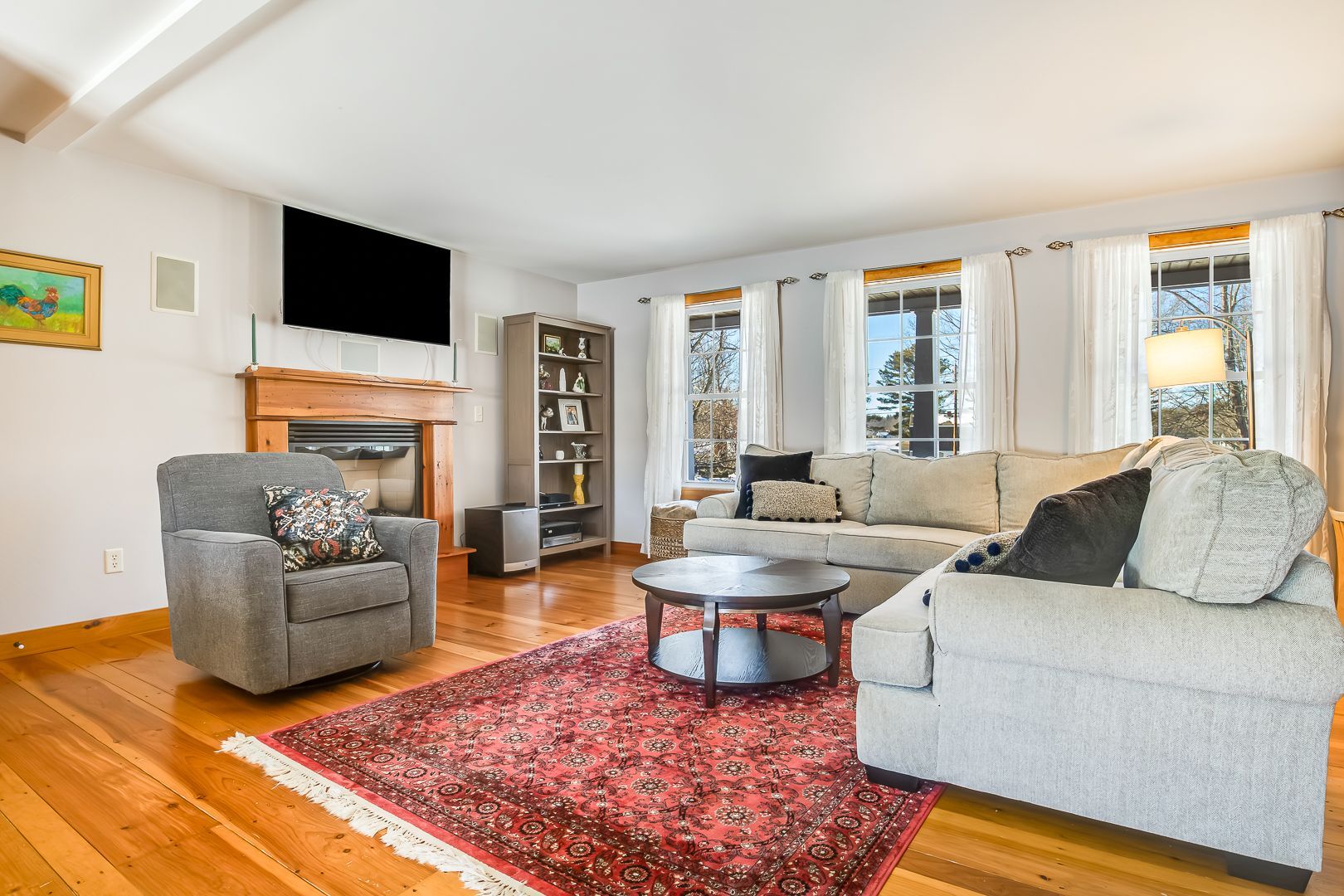 Bright living room with gray sectional, armchair, red rug, hardwood floors, and fireplace with TV above it