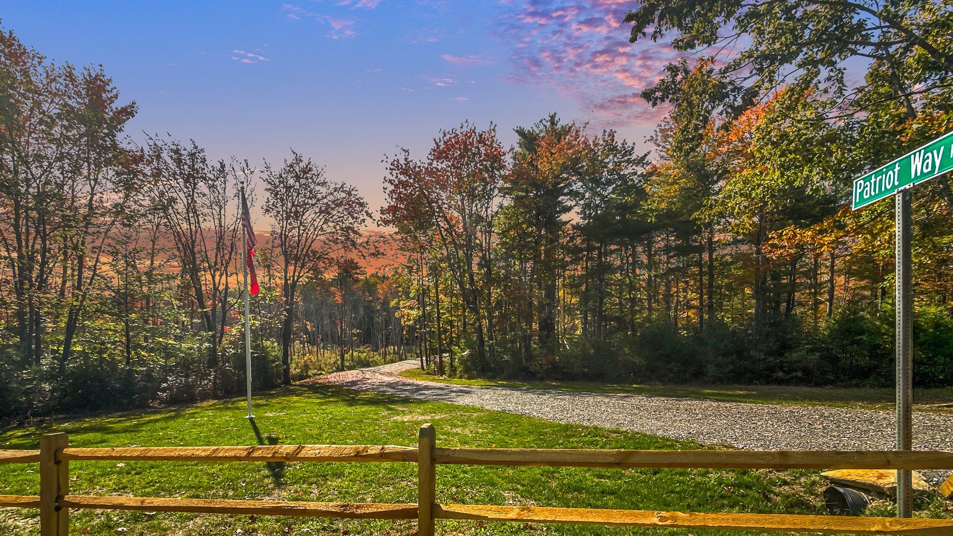 Sunset over a wooded road with a wooden fence, green grass, and a street sign at right