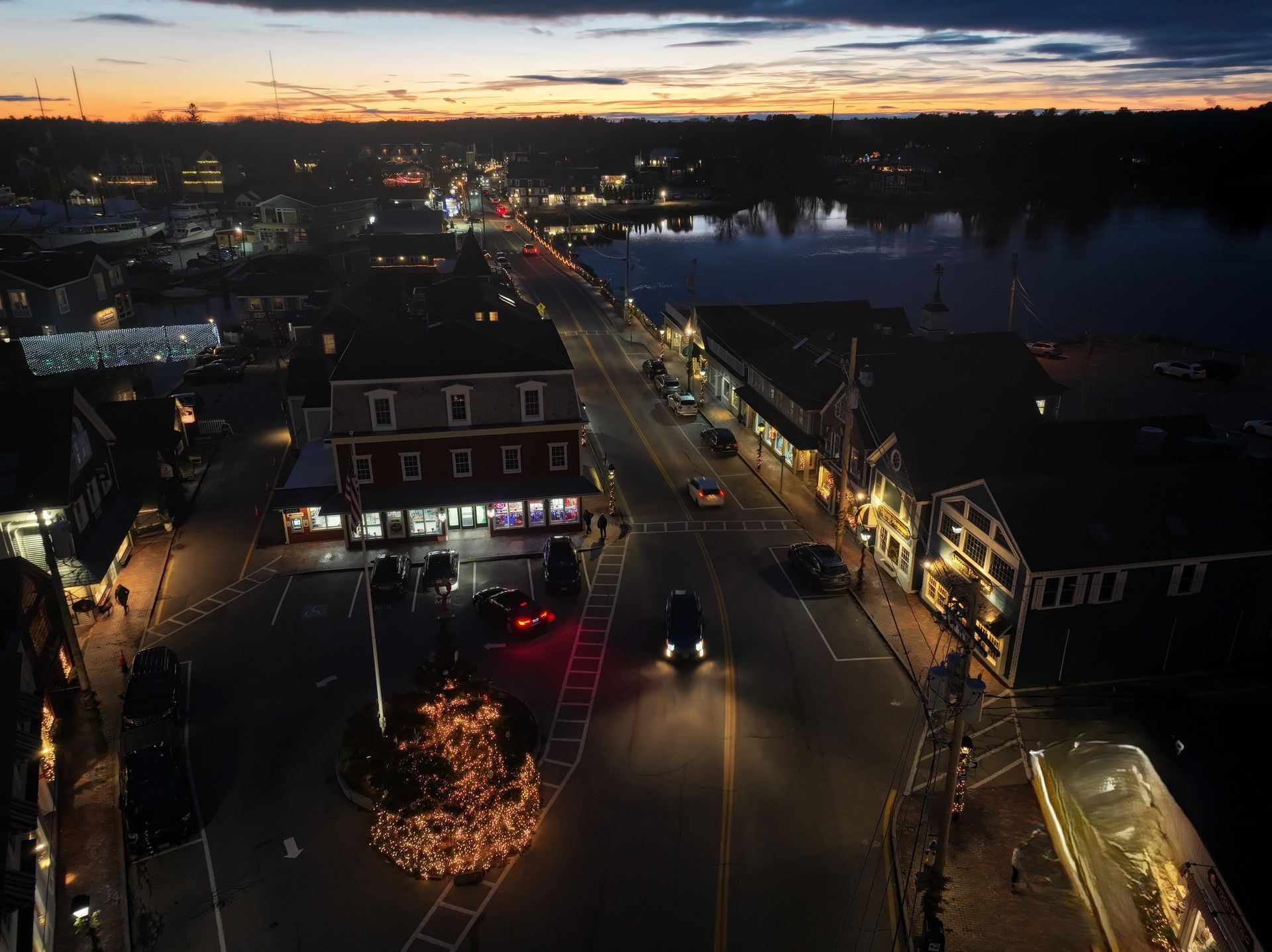 Dusk city street by a river, with lit buildings, traffic, and a glowing tree in the foreground.