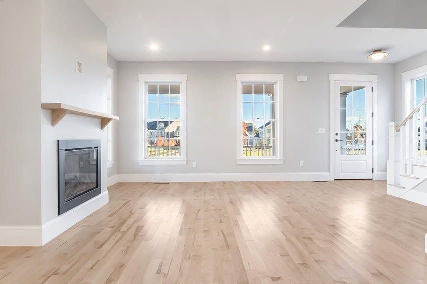 Bright empty living room with hardwood floors, fireplace, and three windows by a front door