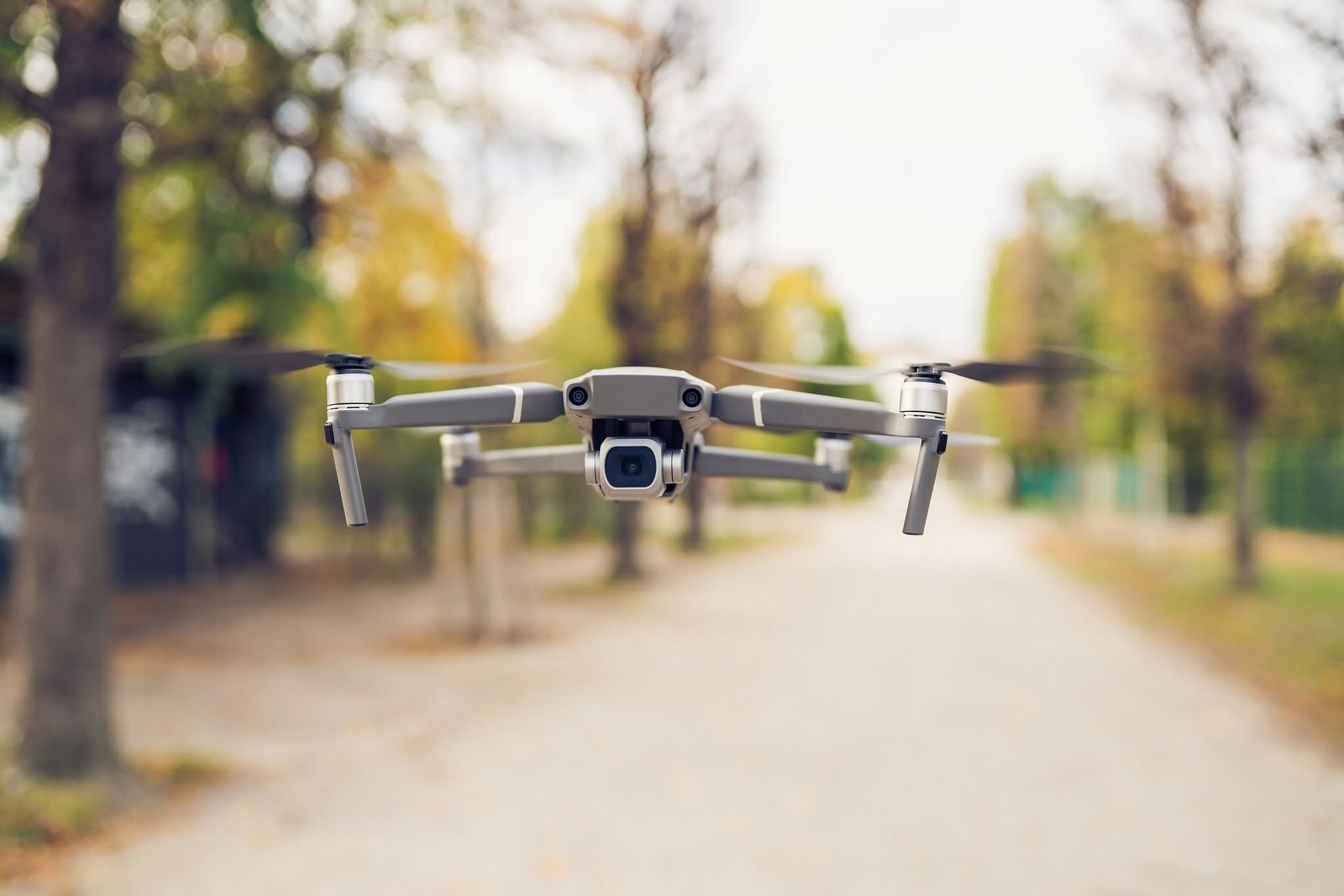 A gray drone hovers on a tree-lined path with blurred autumn background.