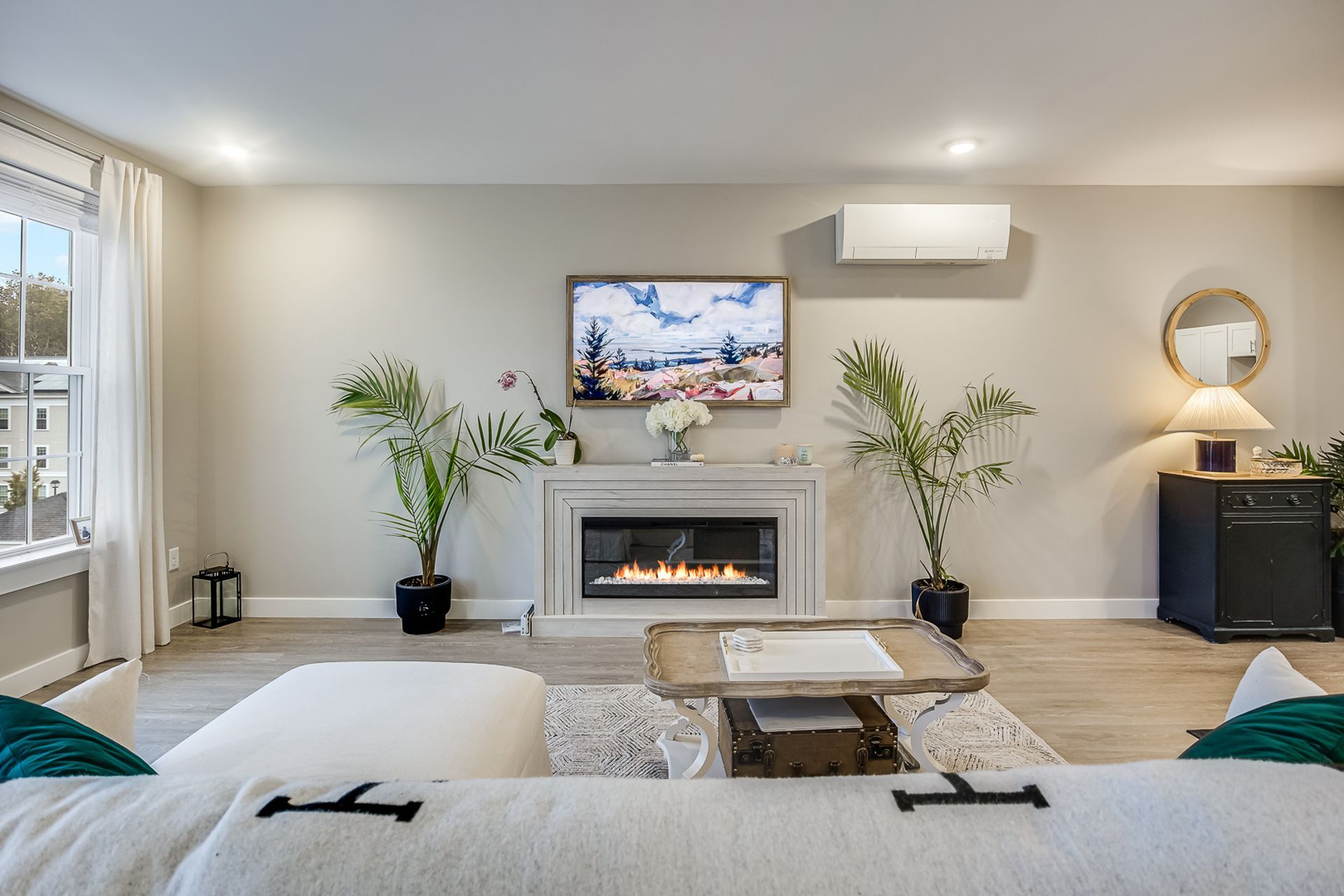 Modern living room with wall-mounted TV, electric fireplace, white sofa, plants, and large window.