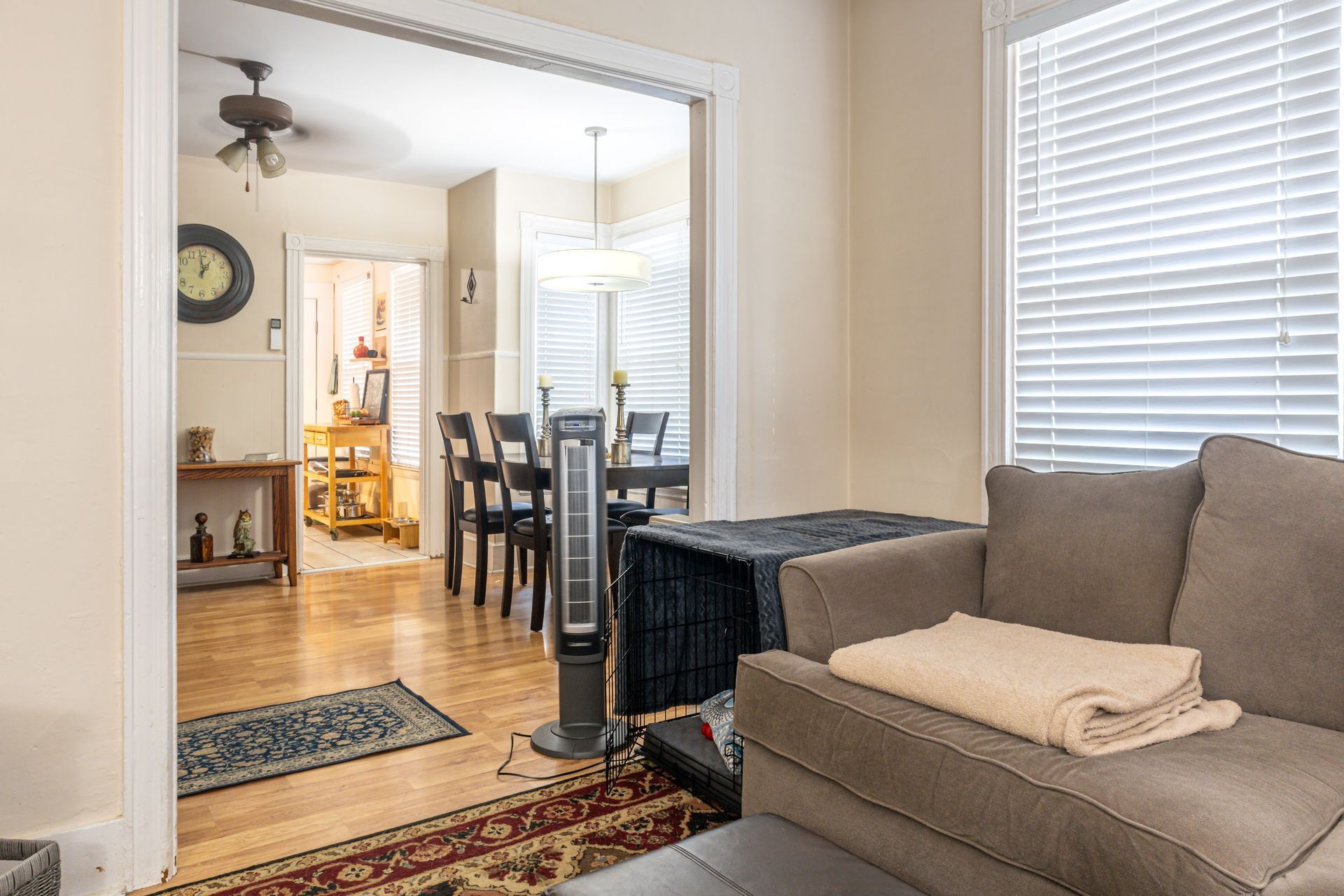 Living room with gray sofa, window blinds, and view into a dining area with dark chairs and hardwood floors