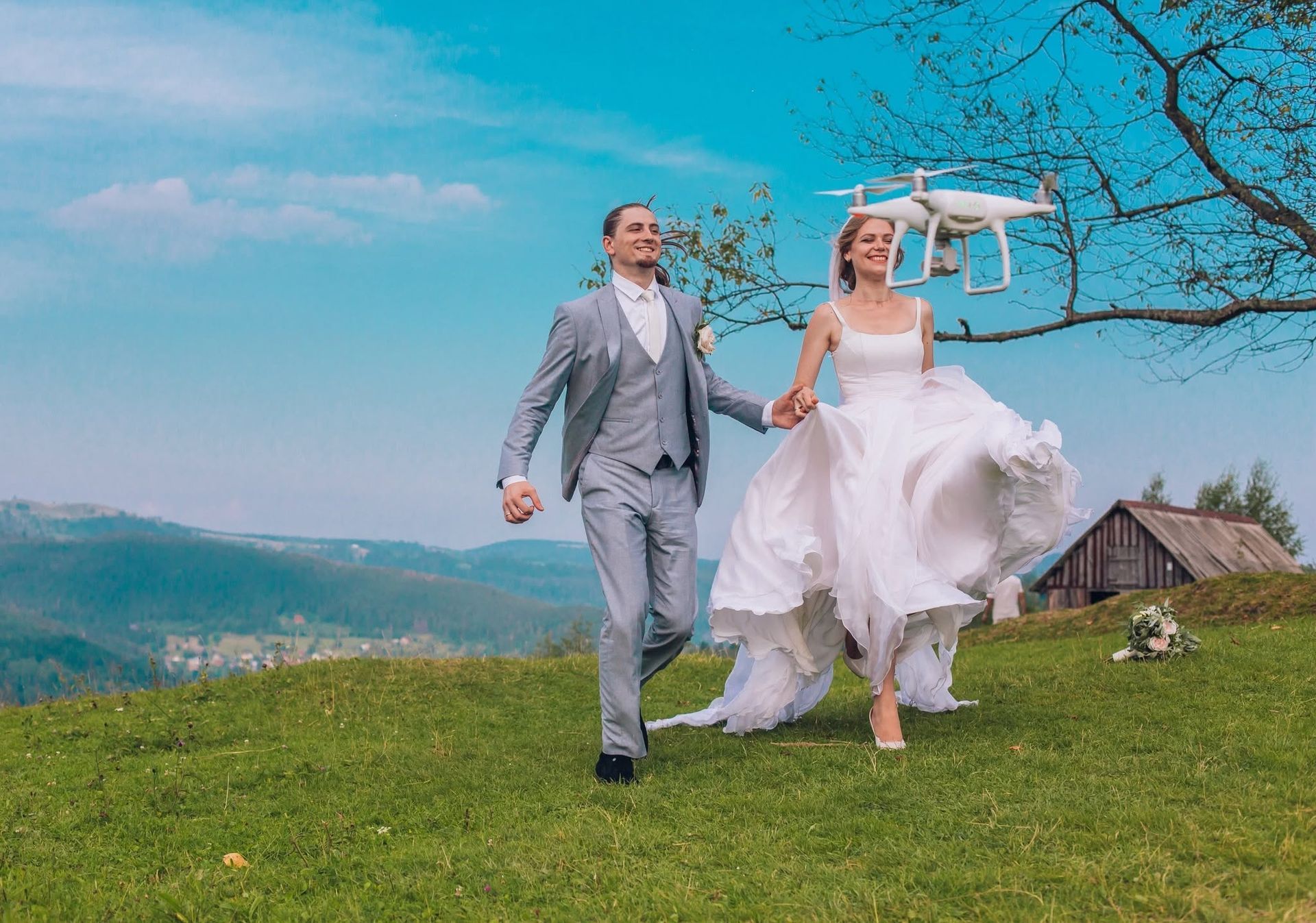 Bride and groom running on a grassy hill with a white drone overhead, under a bright blue sky