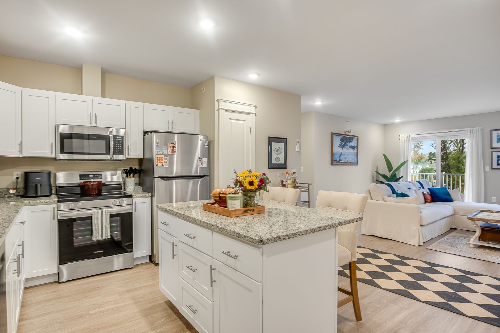 Bright open kitchen and living room with white cabinets, granite island, stainless appliances, and patterned rug