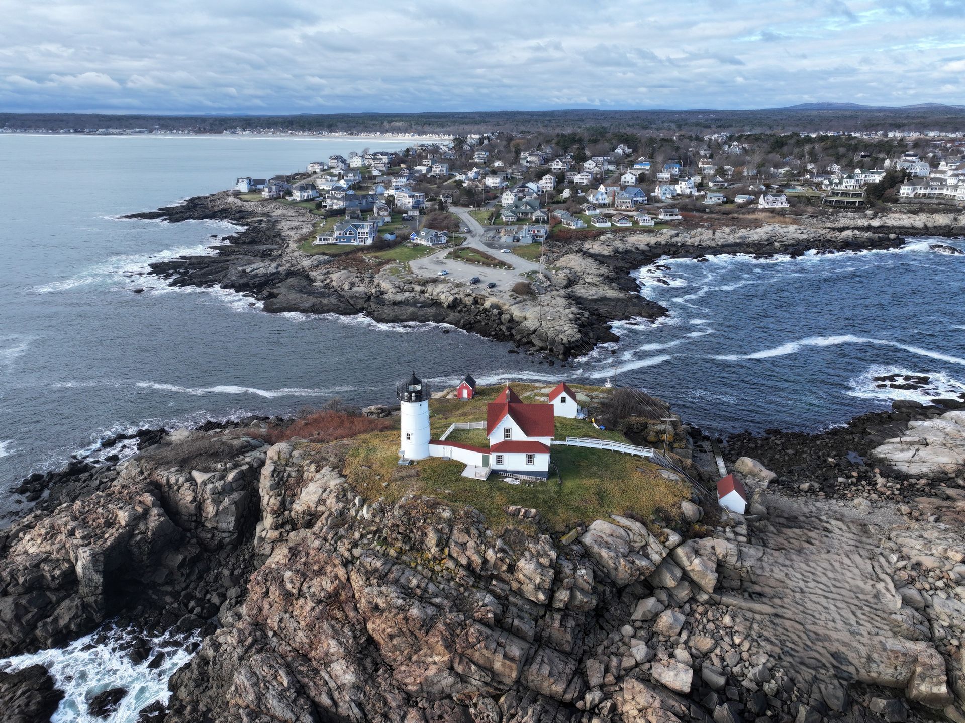 Aerial view of a rocky island lighthouse with a red-roofed keeper’s house and coastal village beyond.