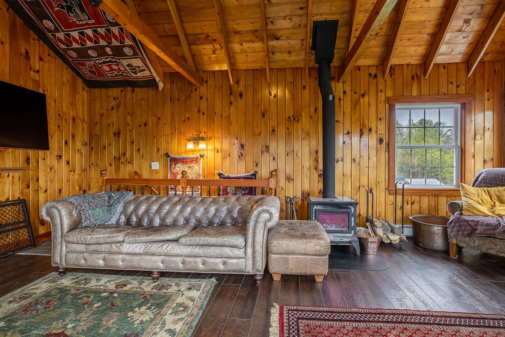 Rustic wood-paneled living room with a gray sofa, wood stove, and large window