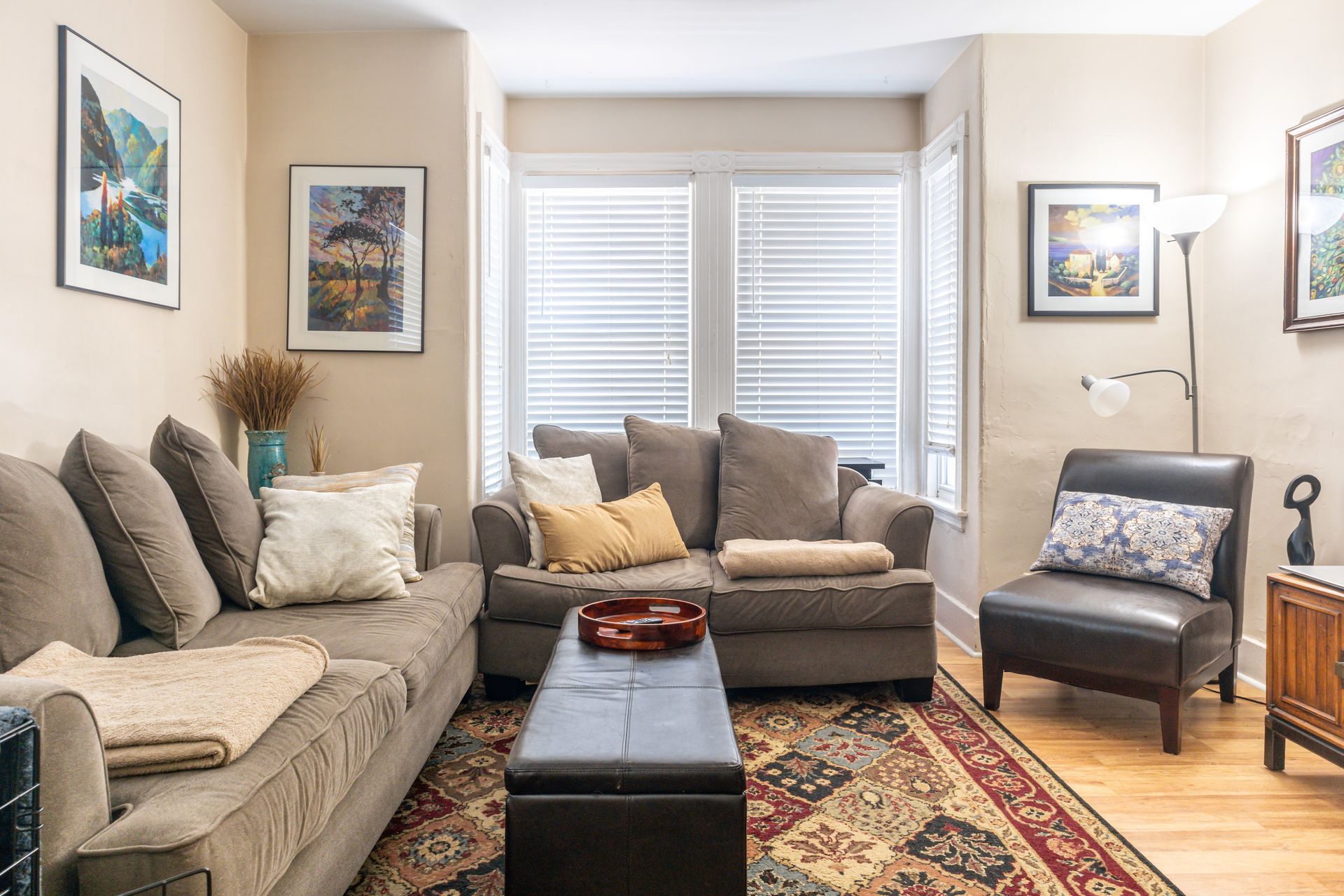 Bright living room with beige sectional, patterned rug, armchair, floor lamp, and shuttered windows
