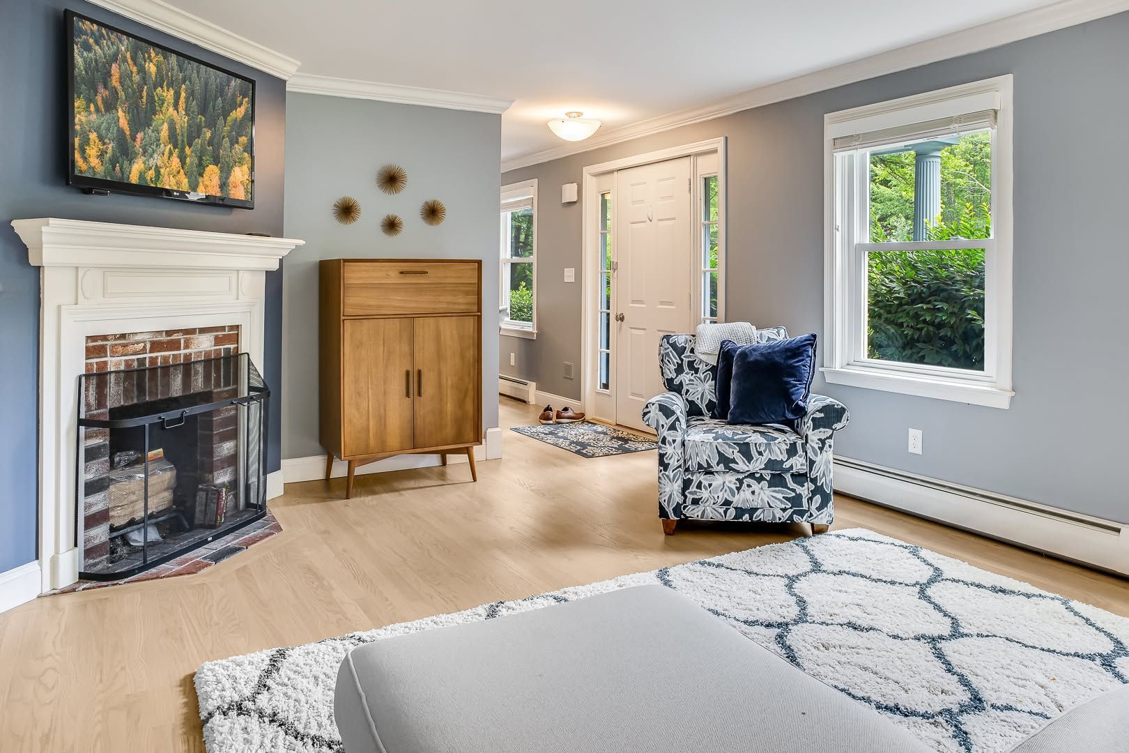 Bright living room with a fireplace, wooden cabinet, patterned chair, and large window with gray walls.