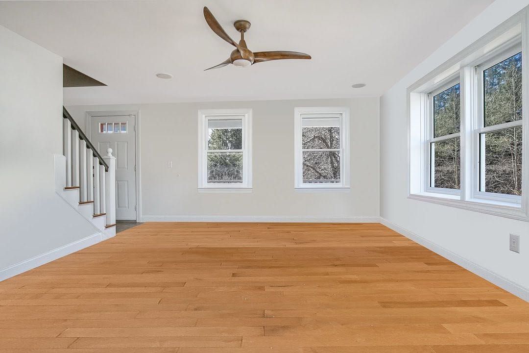 Empty living room with hardwood floors, white walls, ceiling fan, staircase, and large windows.