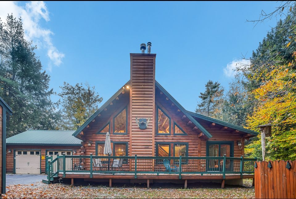 Rustic log cabin with a front porch and stone chimney, surrounded by autumn trees under a blue sky