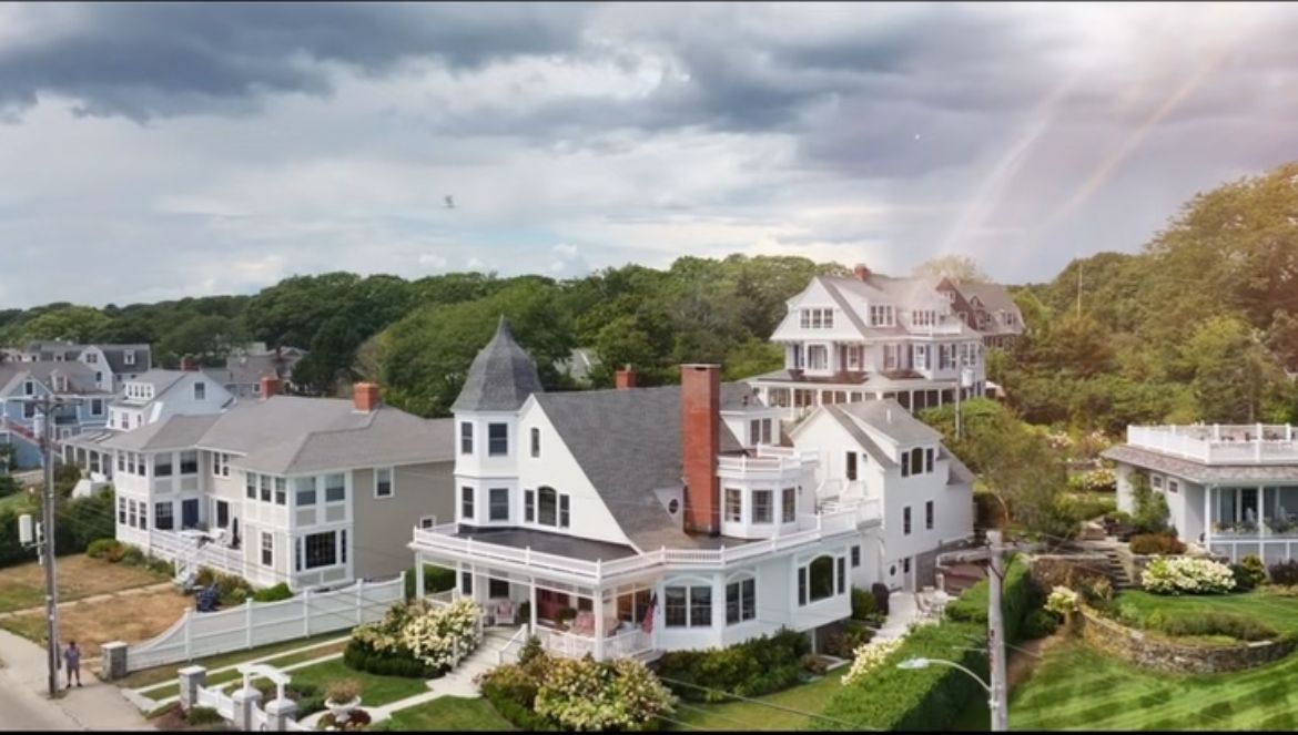 Coastal neighborhood with large white Victorian-style homes and lush green trees under a cloudy sky