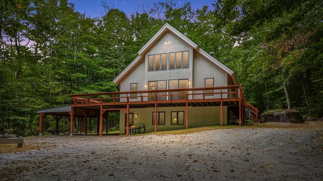 White cabin with a wooden deck in a wooded clearing, surrounded by trees and gravel.