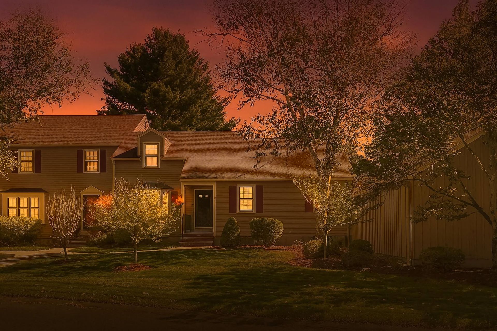 Cozy house at dusk with lit windows, warm orange sky, and trees framing the yard