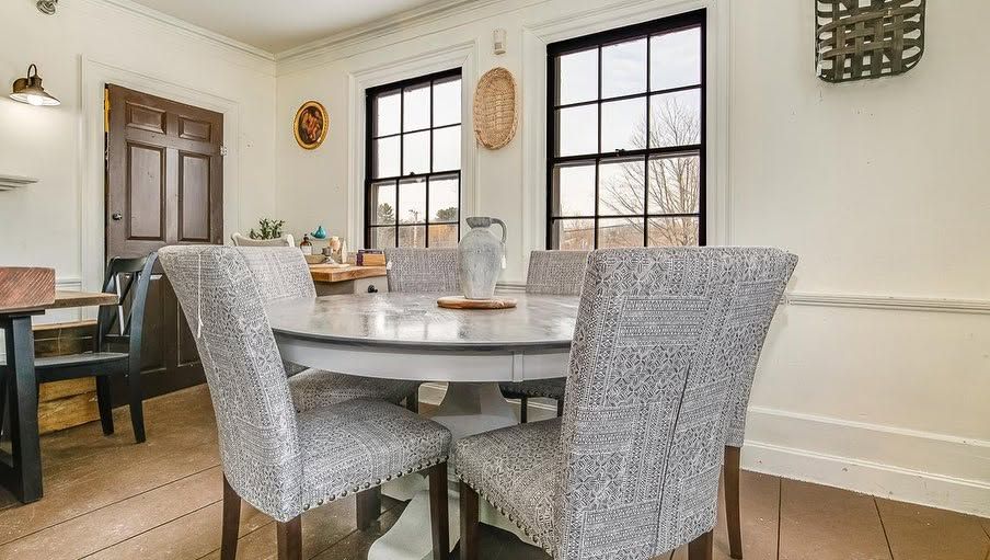 Bright dining room with a round table, gray chairs, windows, and a wooden cabinet.