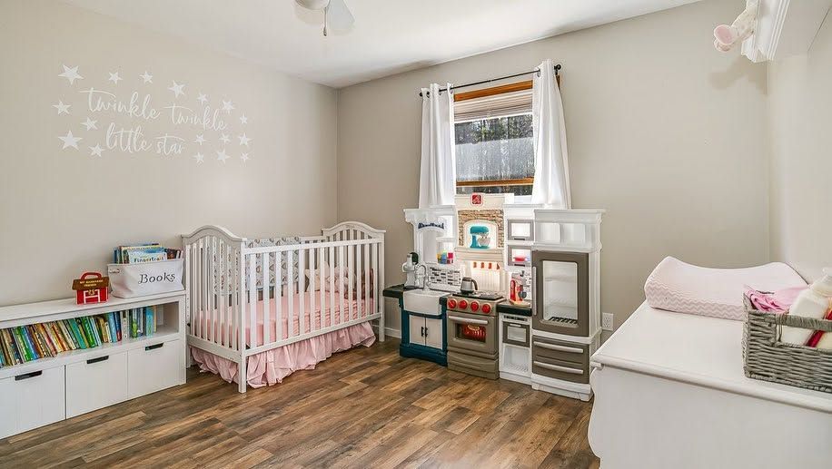 Bright nursery with two white cribs, toy shelves, a window, and a changing table on wood floors.