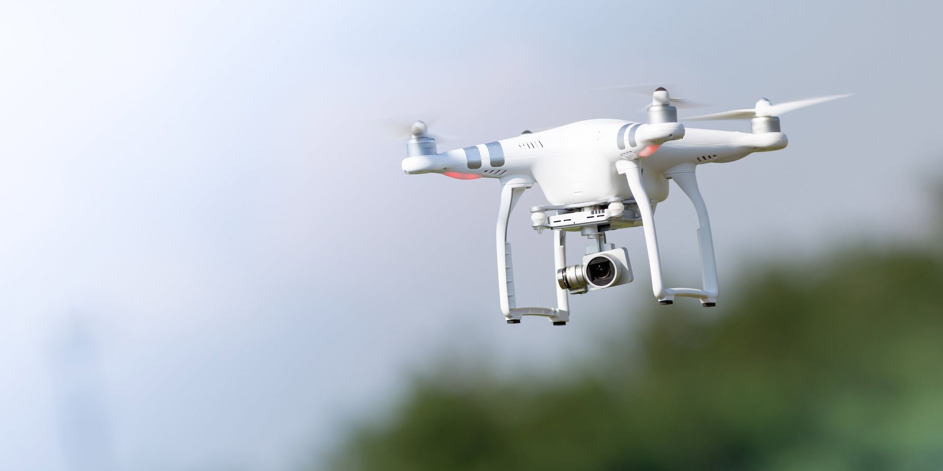 White quadcopter drone flying outdoors against a blurry blue sky and green background