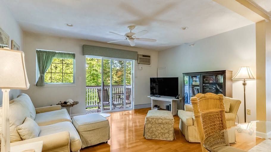 Bright living room with white furniture, wicker chairs, TV, and glass doors to greenery