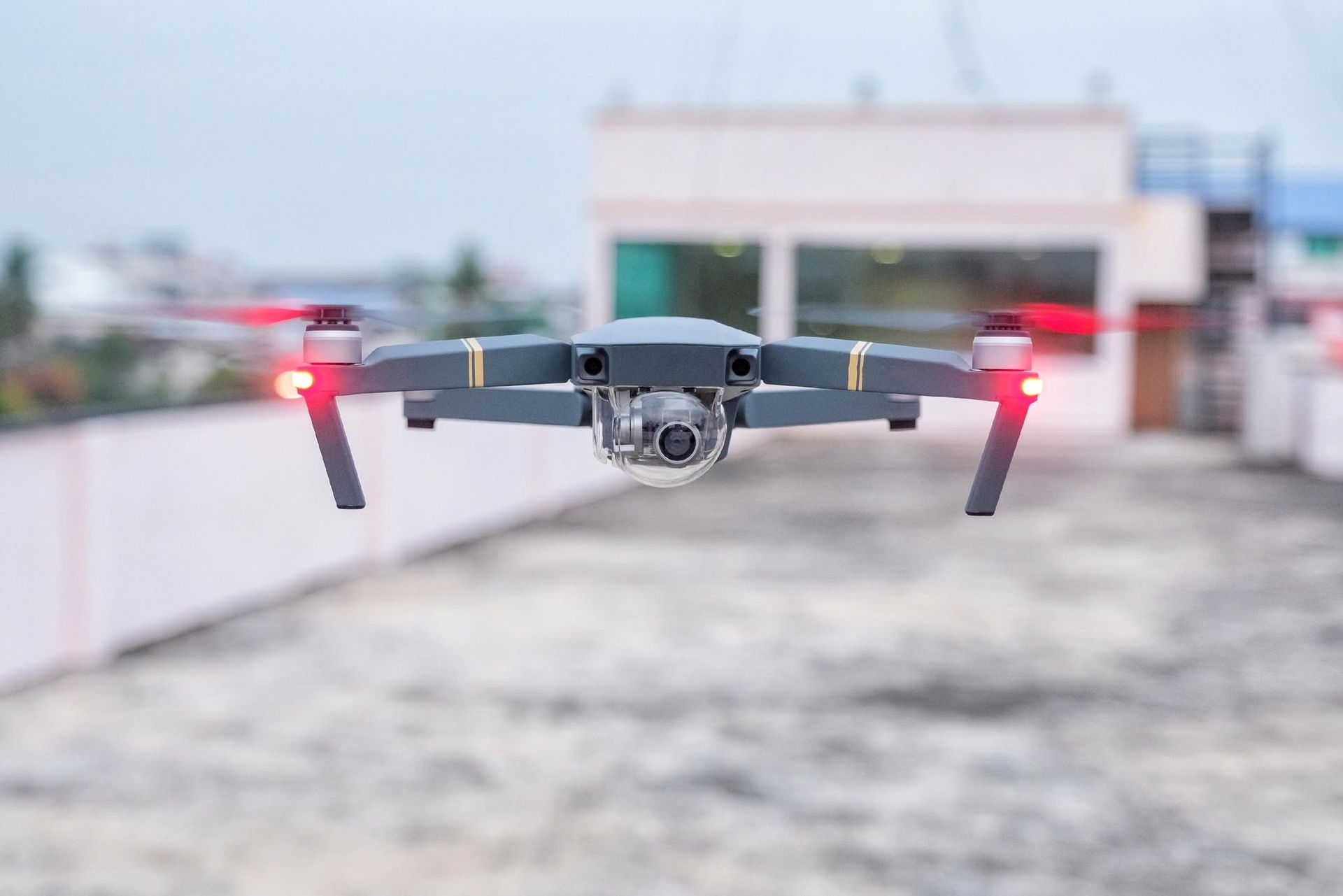 Gray drone hovering outdoors with red lights on, centered against a blurred rooftop background