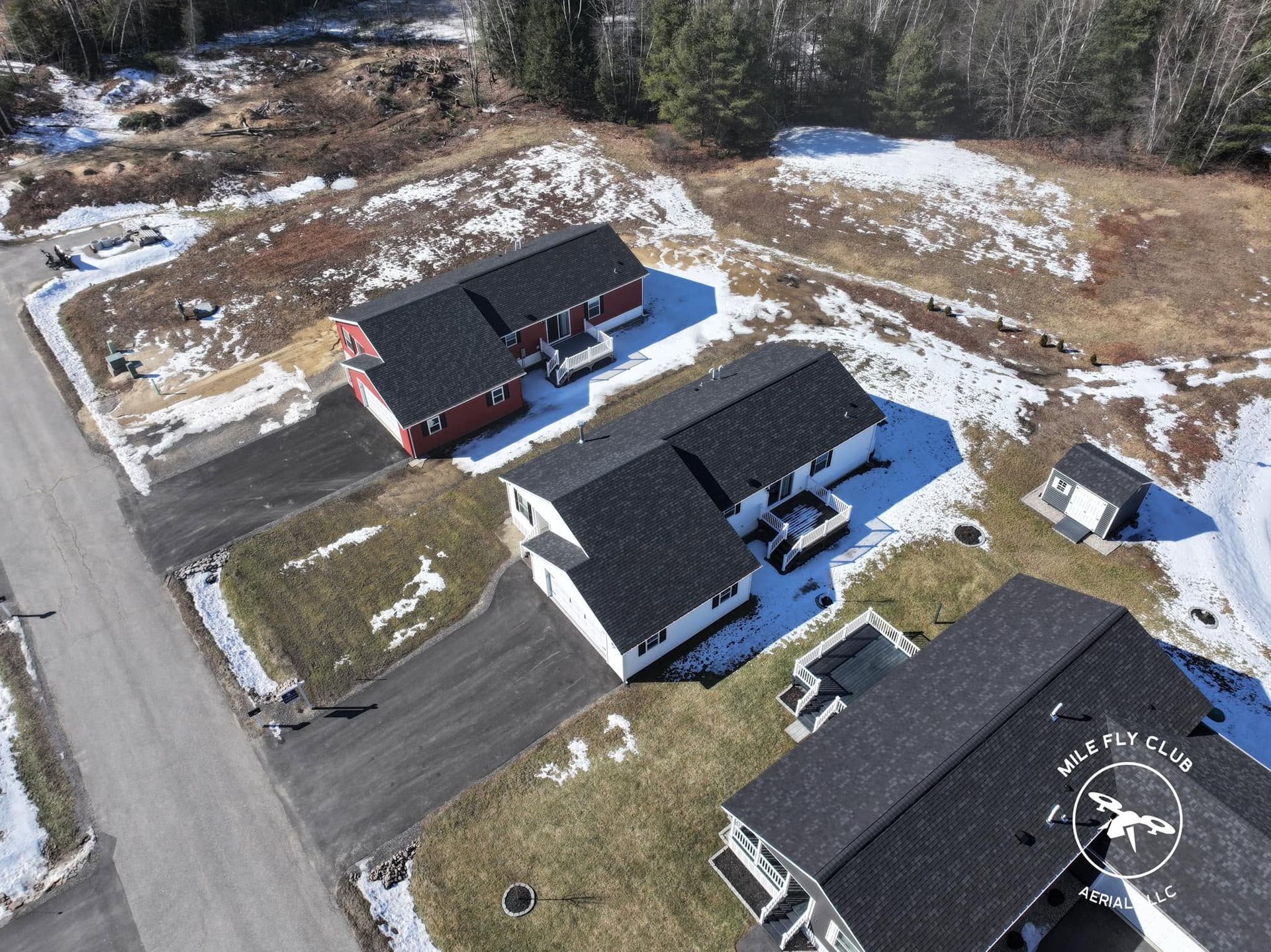 Aerial view of three dark-roofed houses and driveways in a snowy suburban neighborhood with woods nearby