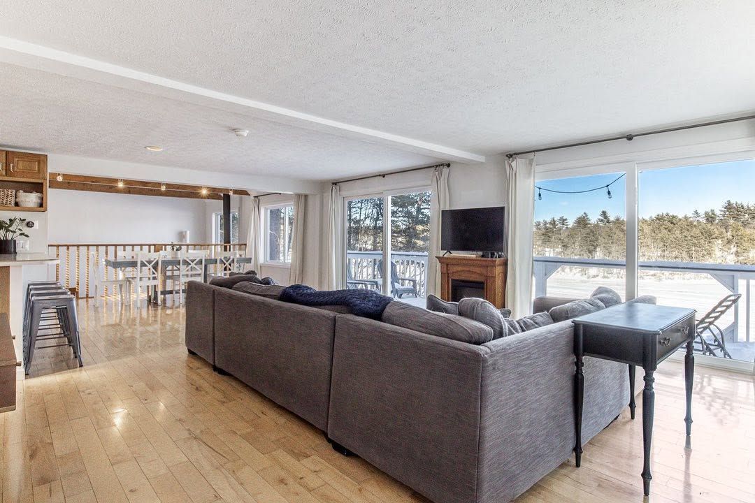 Bright living room with gray sectional sofa, wooden floors, fireplace, and large windows overlooking water.