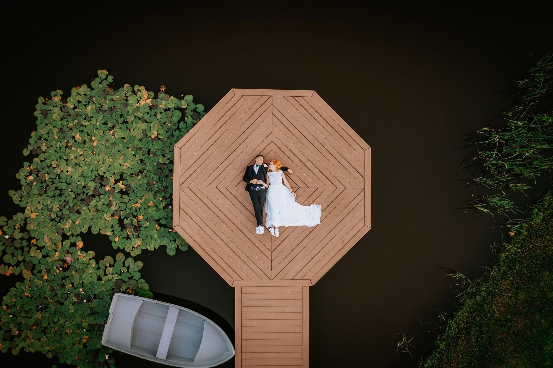 Couple lying on a wooden dock over dark water, with a small boat and trees nearby.
