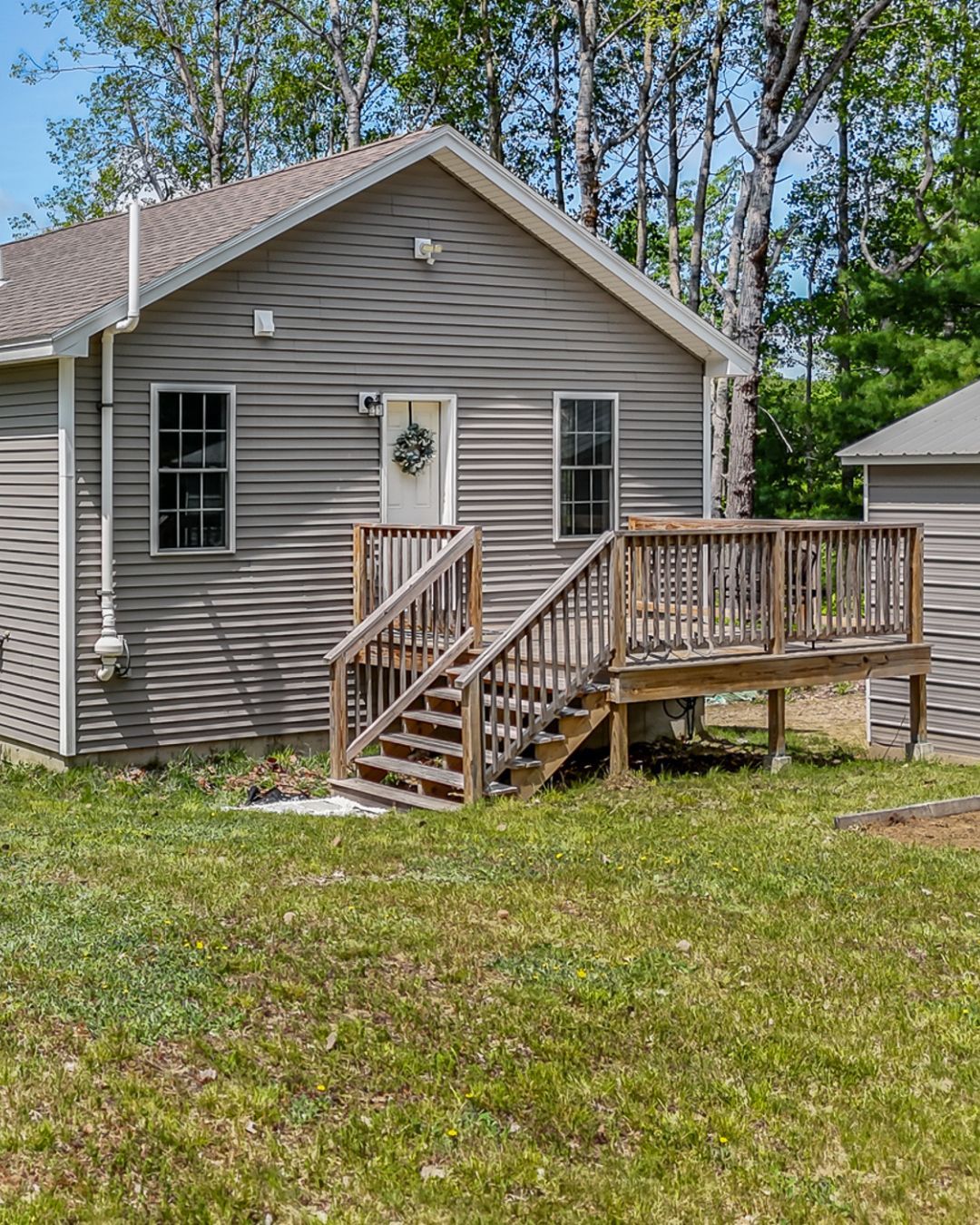 Gray house with a wooden ramp and steps, white door, and grassy yard with trees in back.