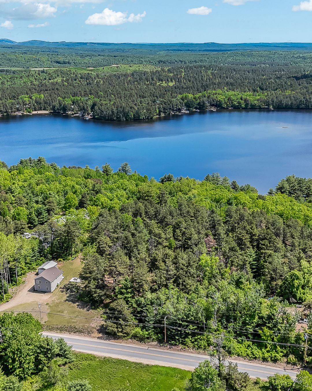 Aerial view of a blue lake bordered by dense green forest, with a road and small house in the foreground