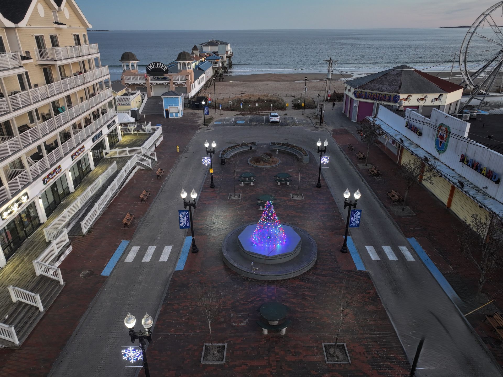 Seaside plaza with a glowing purple fountain at dusk, flanked by buildings and lampposts, with the ocean beyond