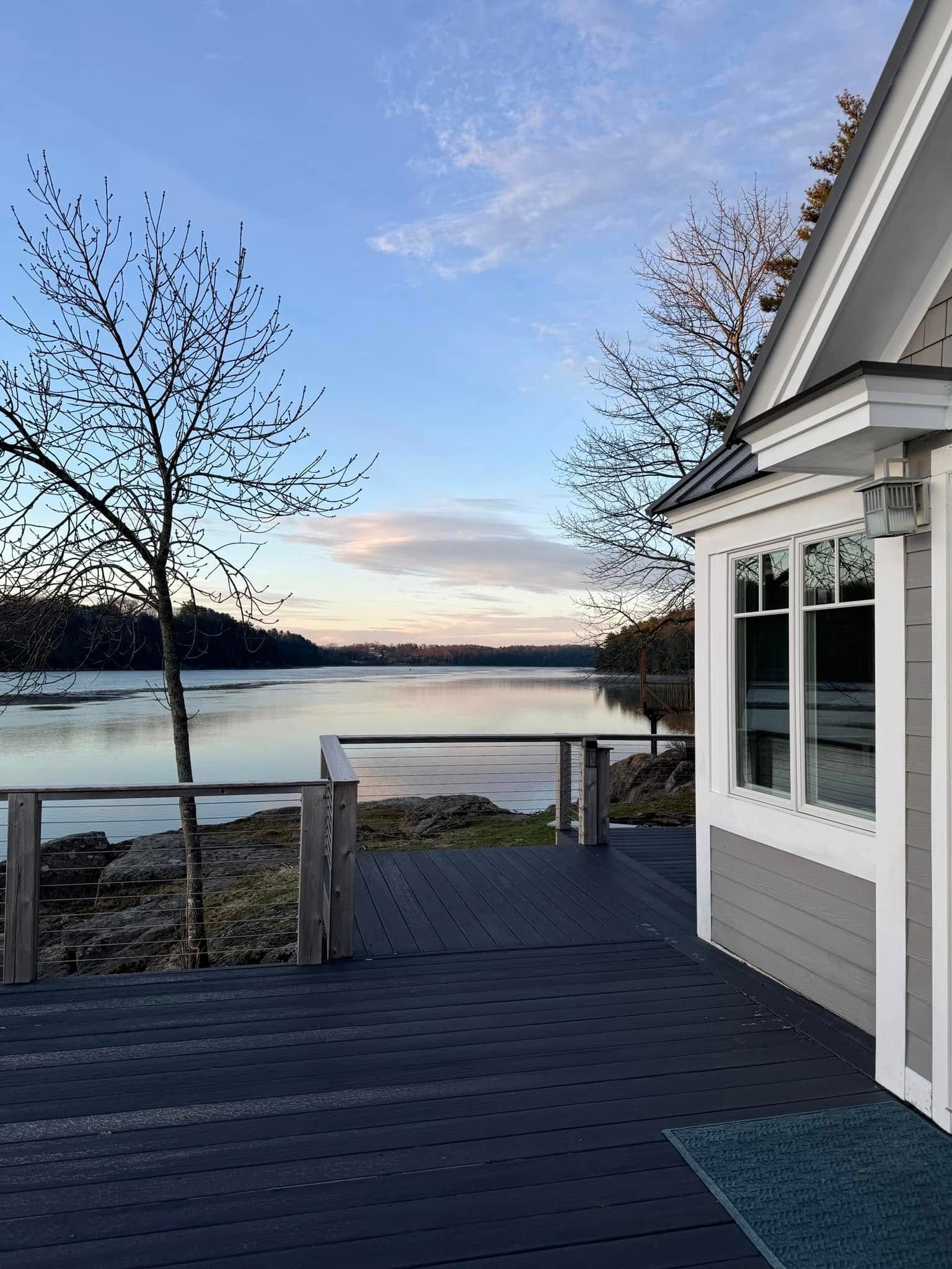 Lakefront deck beside a white house at dusk, with calm water, bare trees, and a pastel sky.