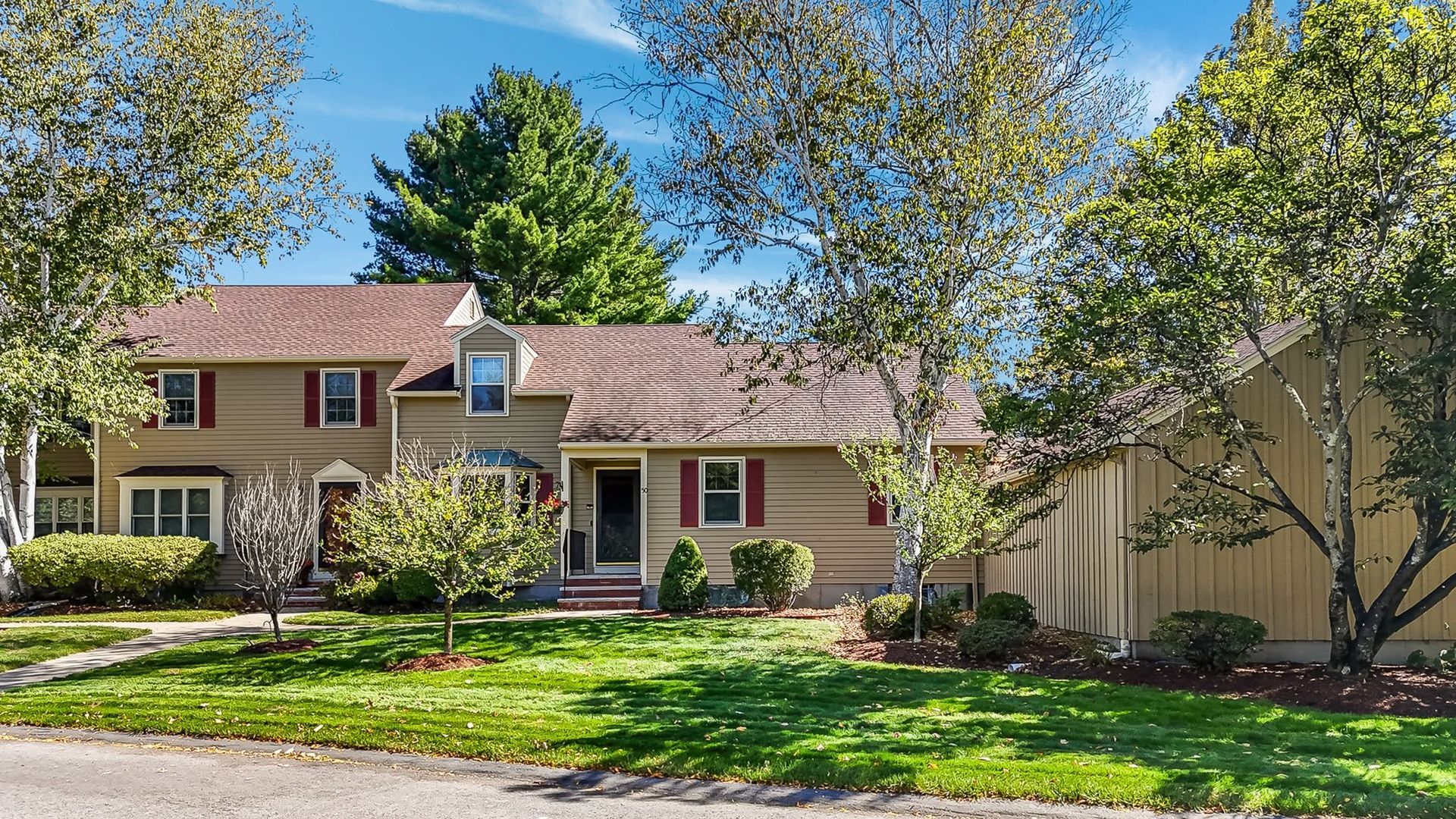 Suburban house with beige siding, red roof, green lawn, and tall trees under a bright blue sky