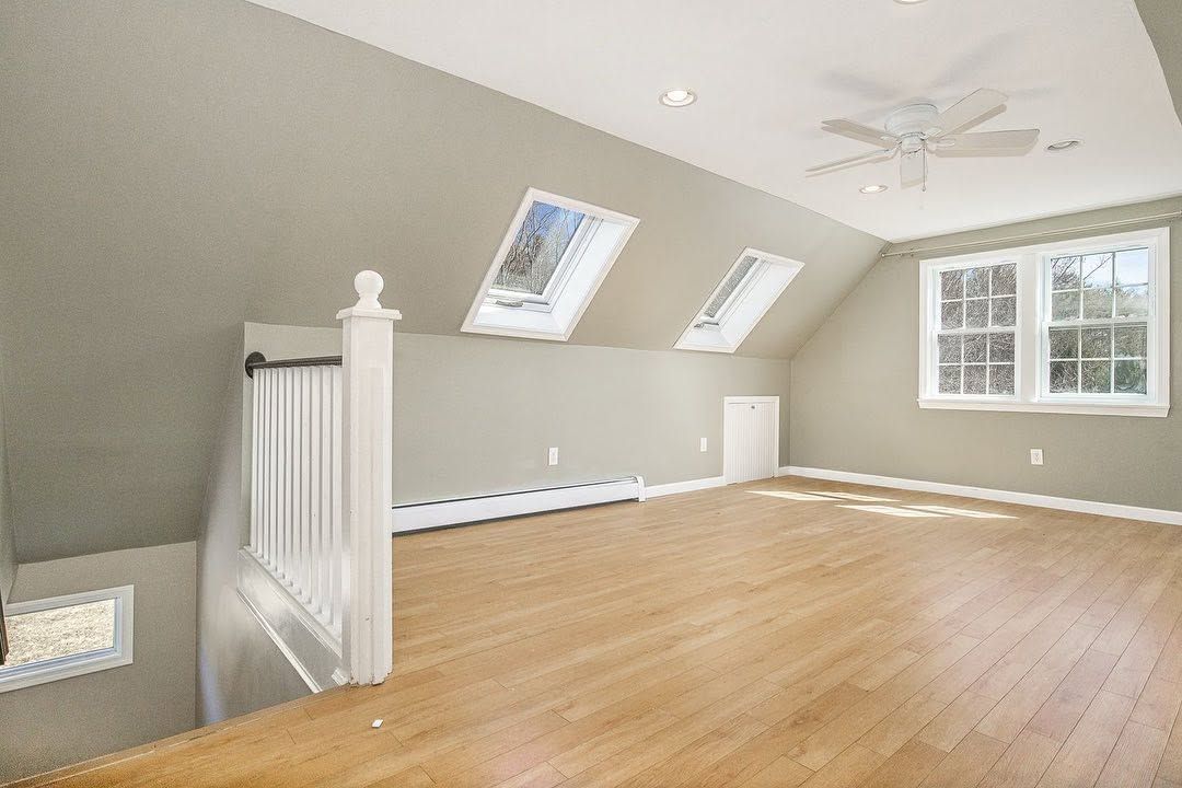 Bright empty attic room with hardwood floor, sloped ceiling, skylights, and a window overlooking stairs