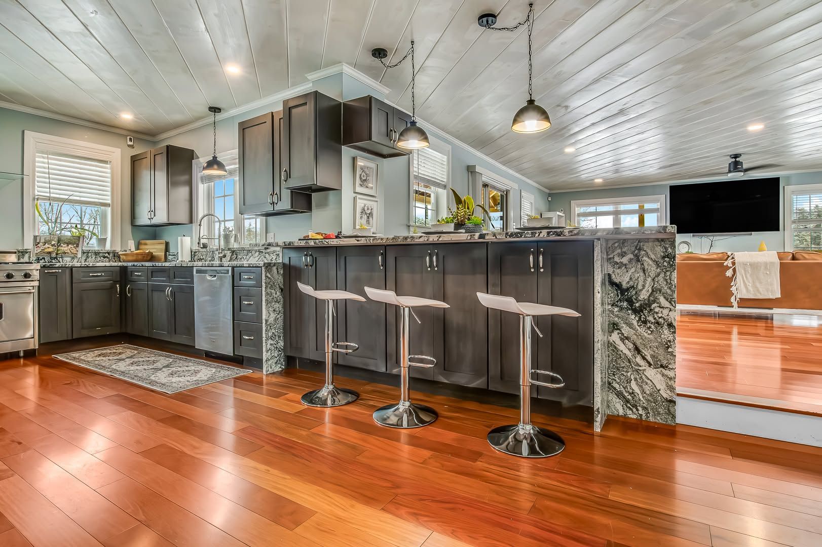 Modern kitchen with a long dark island, bar stools, stainless appliances, and warm wood floors