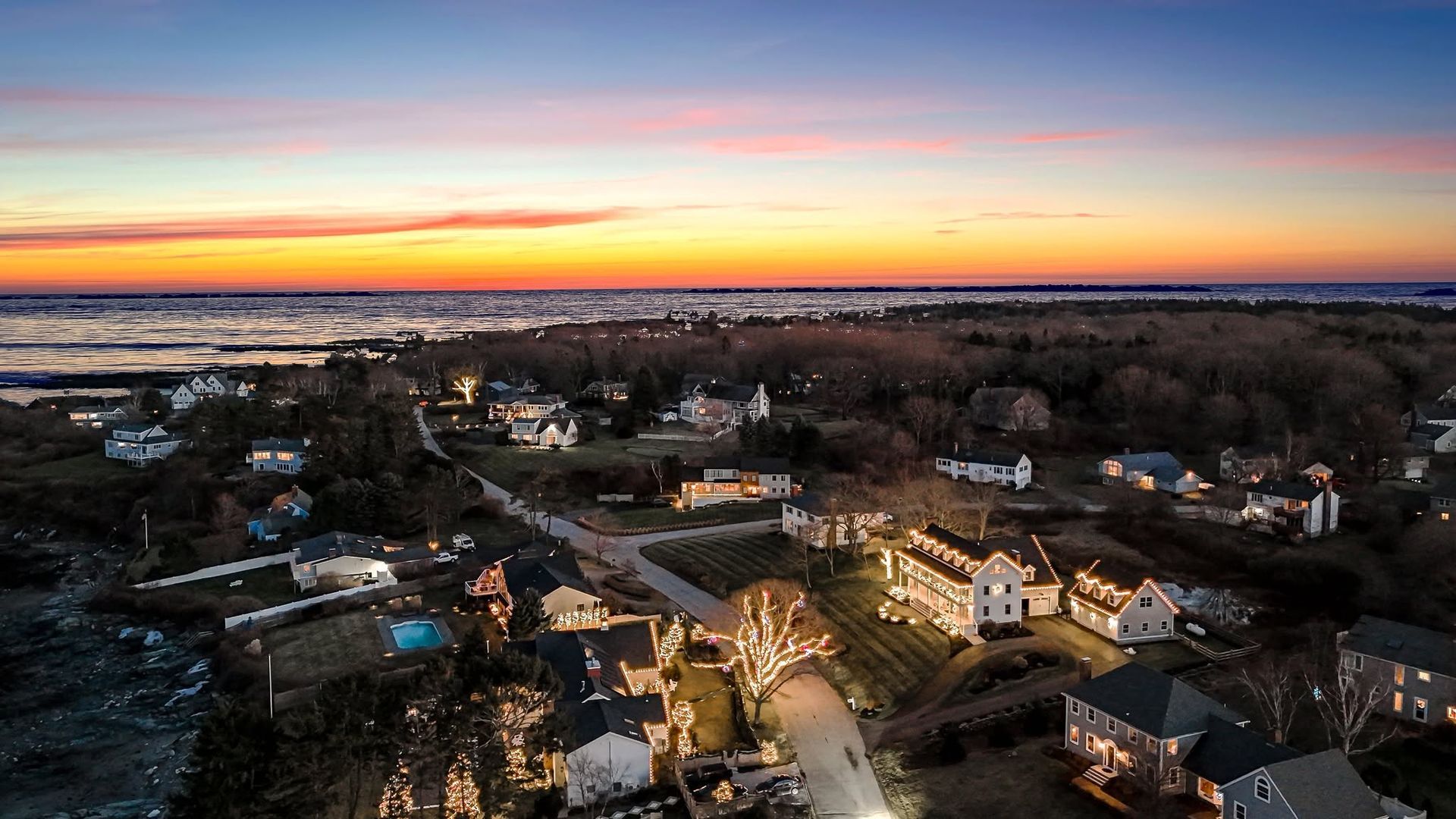 Aerial view of a suburban neighborhood at sunset with glowing streetlights and a colorful horizon.