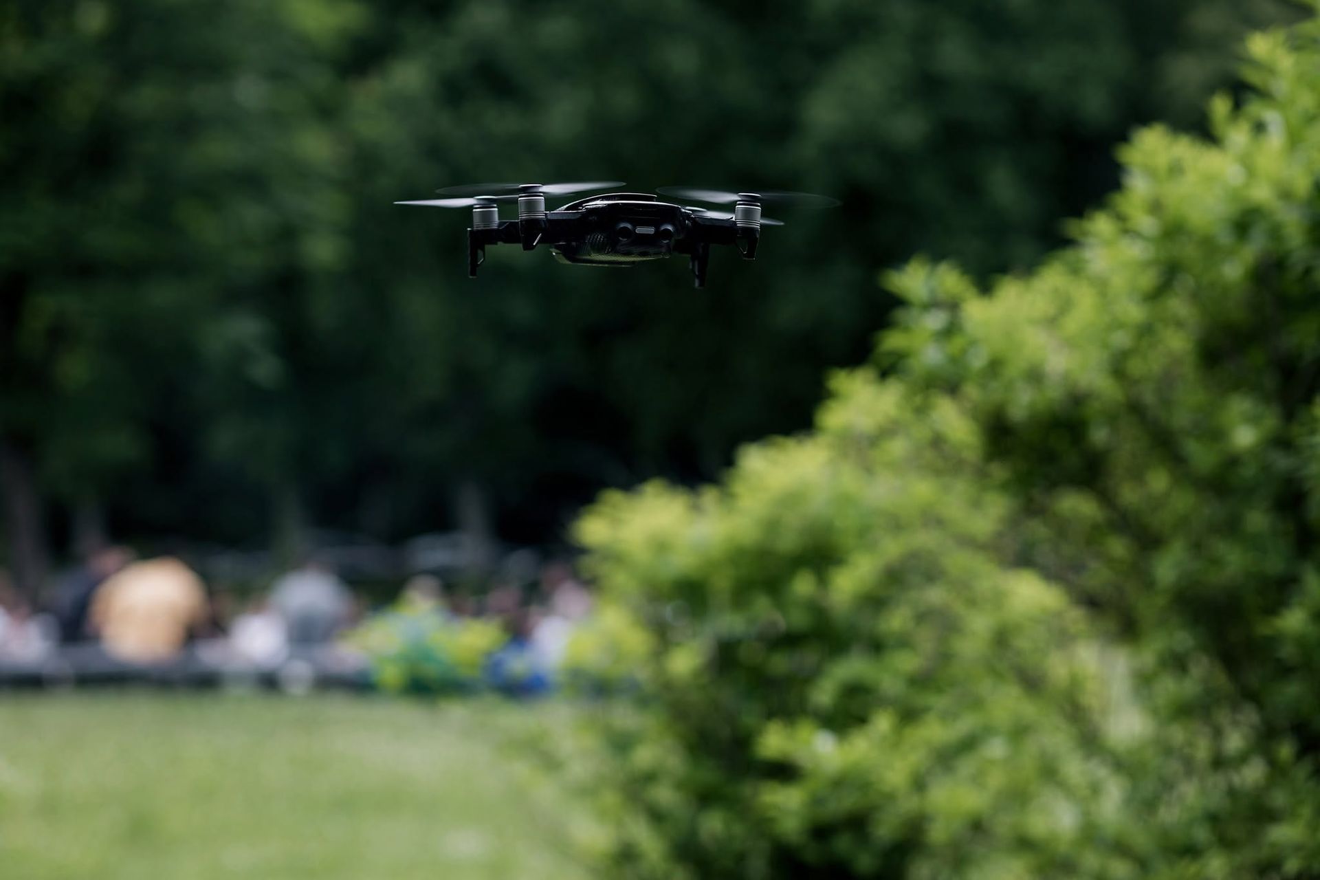 Black drone hovering above a green park with blurred trees in the background