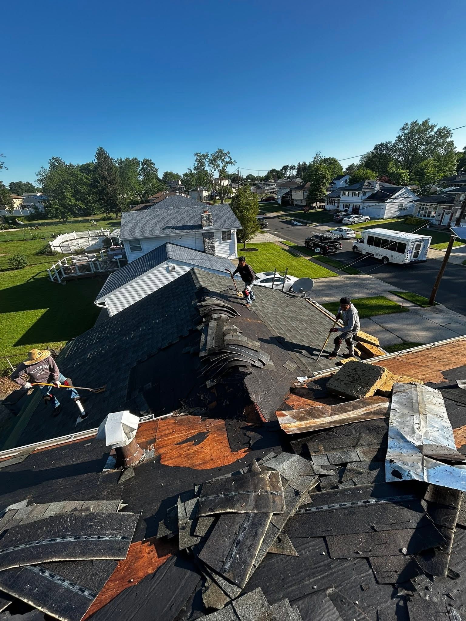 Damaged roof and debris after storm, with suburban houses and a clear blue sky in the background