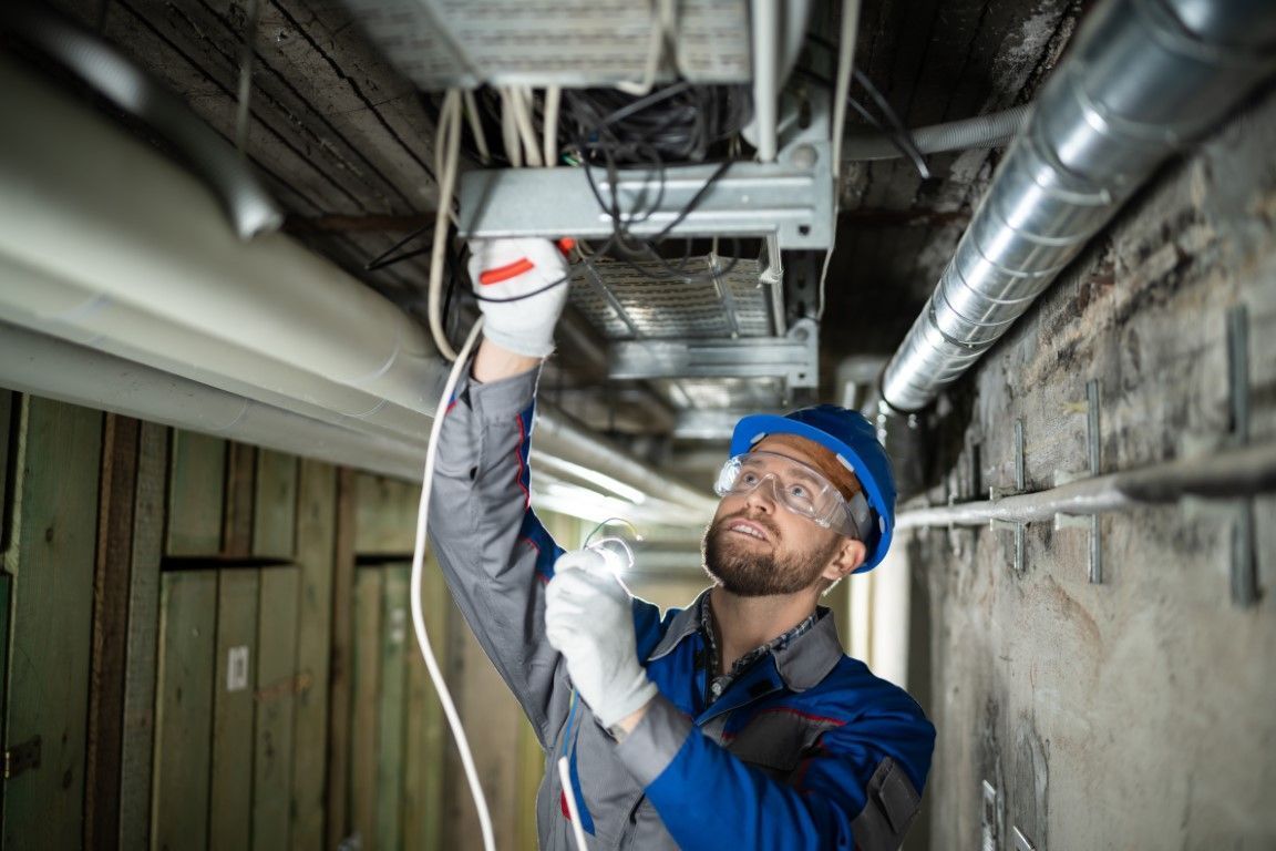 Electrician in blue uniform inspecting ceiling wiring in a narrow utility corridor