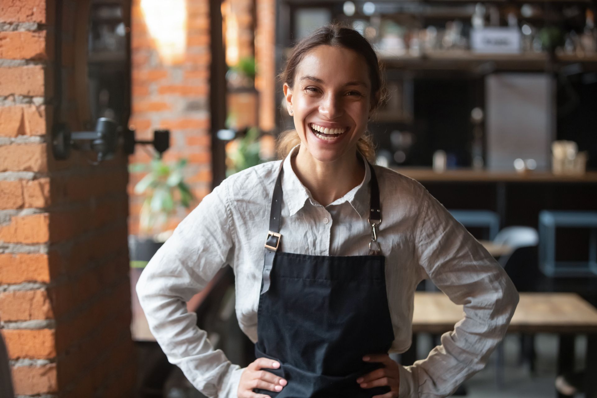 Woman in a black apron smiles, hands on hips, in a restaurant with brick walls.