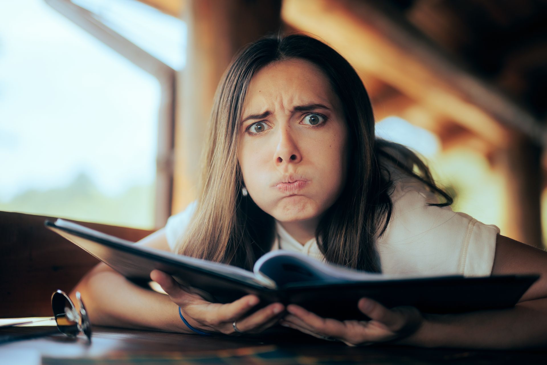 A person with a frustrated expression and puffed cheeks holds an open book while sitting at a table.