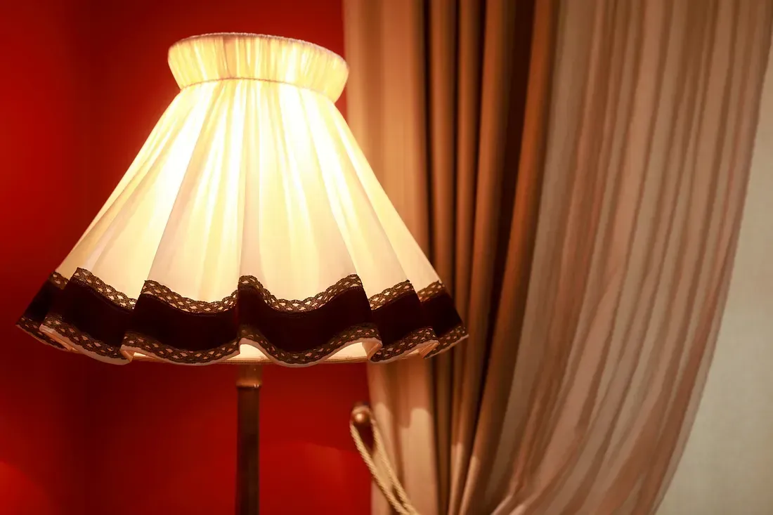 Floor lamp with a cream-colored shade edged with dark lace against a red wall and tan curtains.